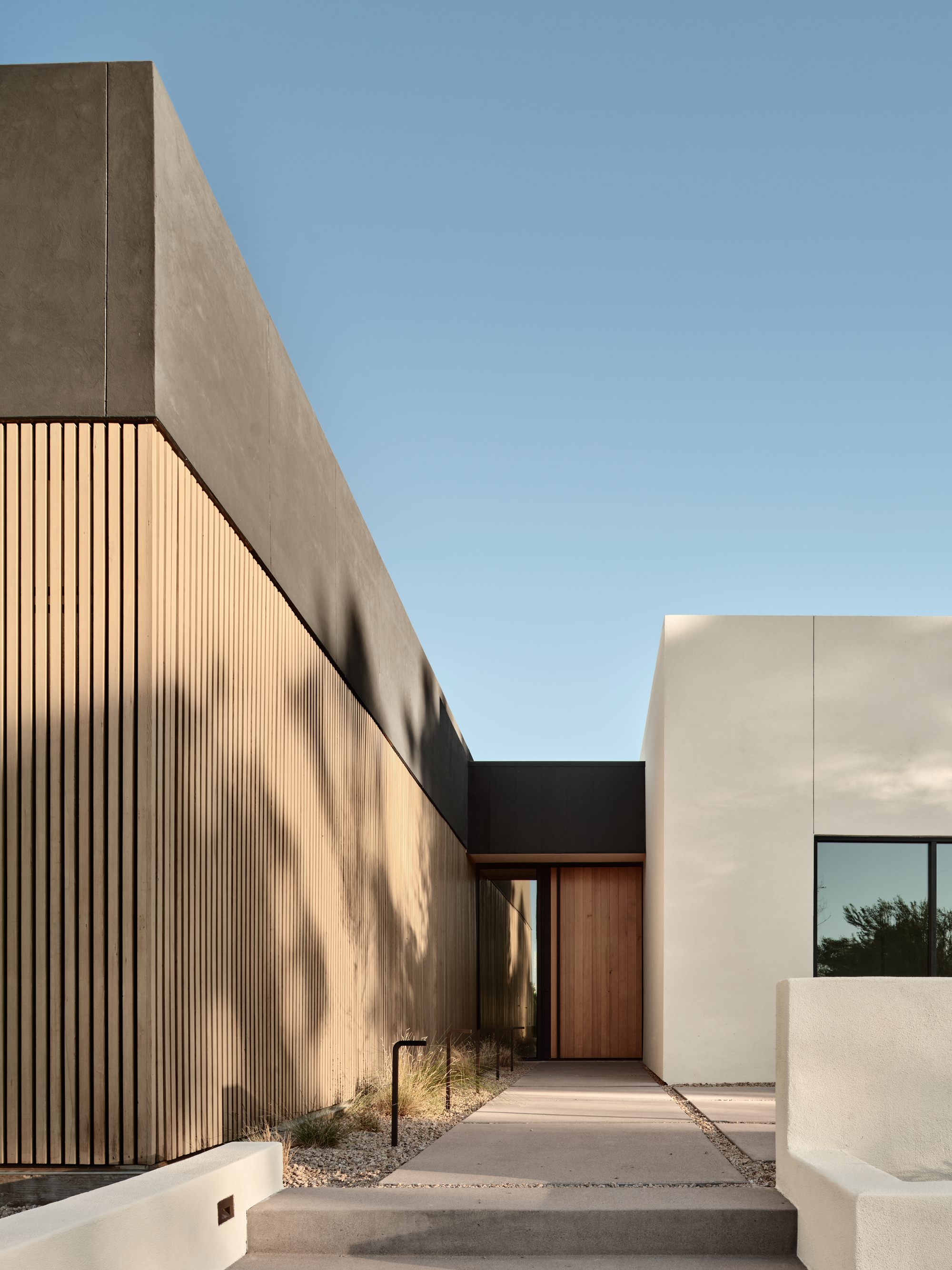 Entry walkway lined by vertical wood slat wall and concrete volumes casting afternoon shadows