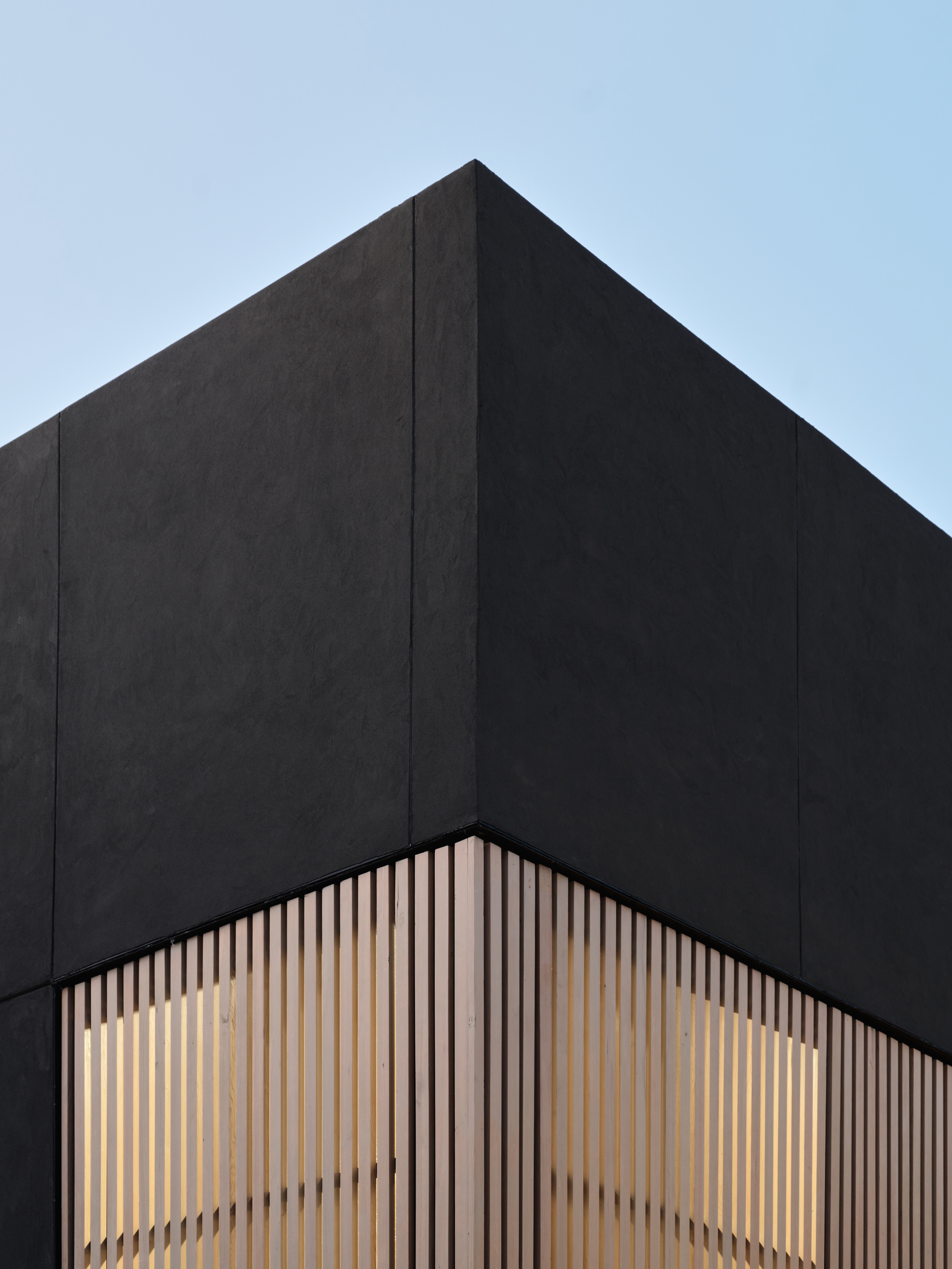 Corner detail of black cladding meeting illuminated timber slat screen at dusk