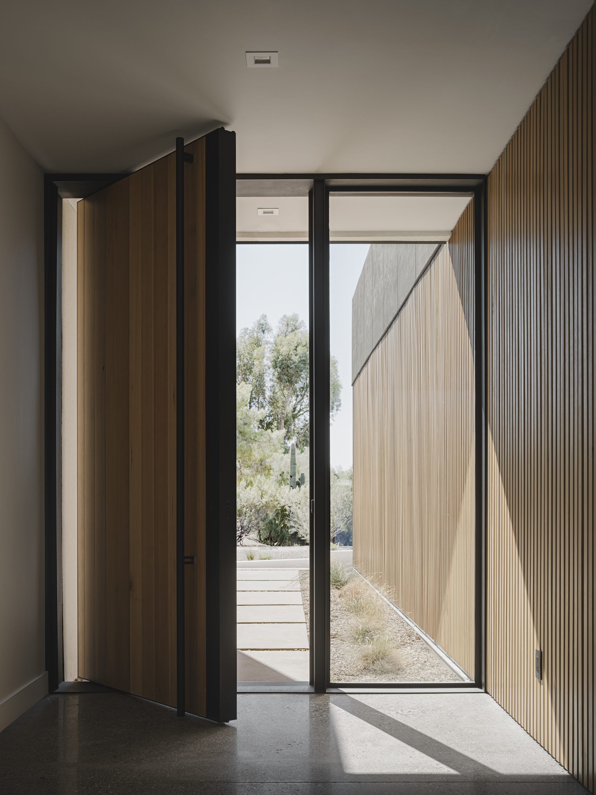 Entry vestibule with pivoting timber door and vertical fluted wood wall panel casting shadows on polished concrete floor