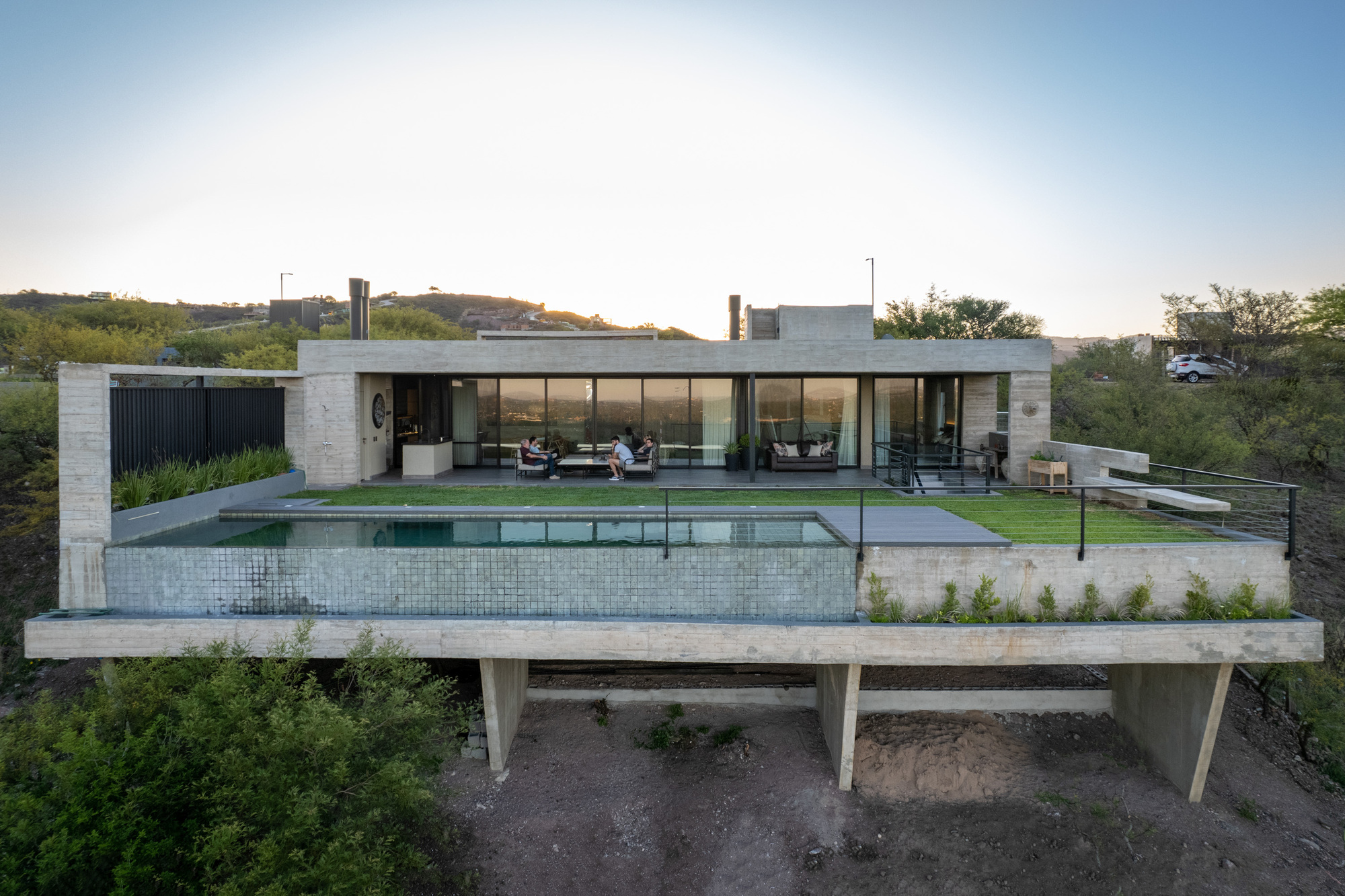 Elevated concrete platform supporting pool terrace and glass-walled pavilion above sloping natural terrain at dusk