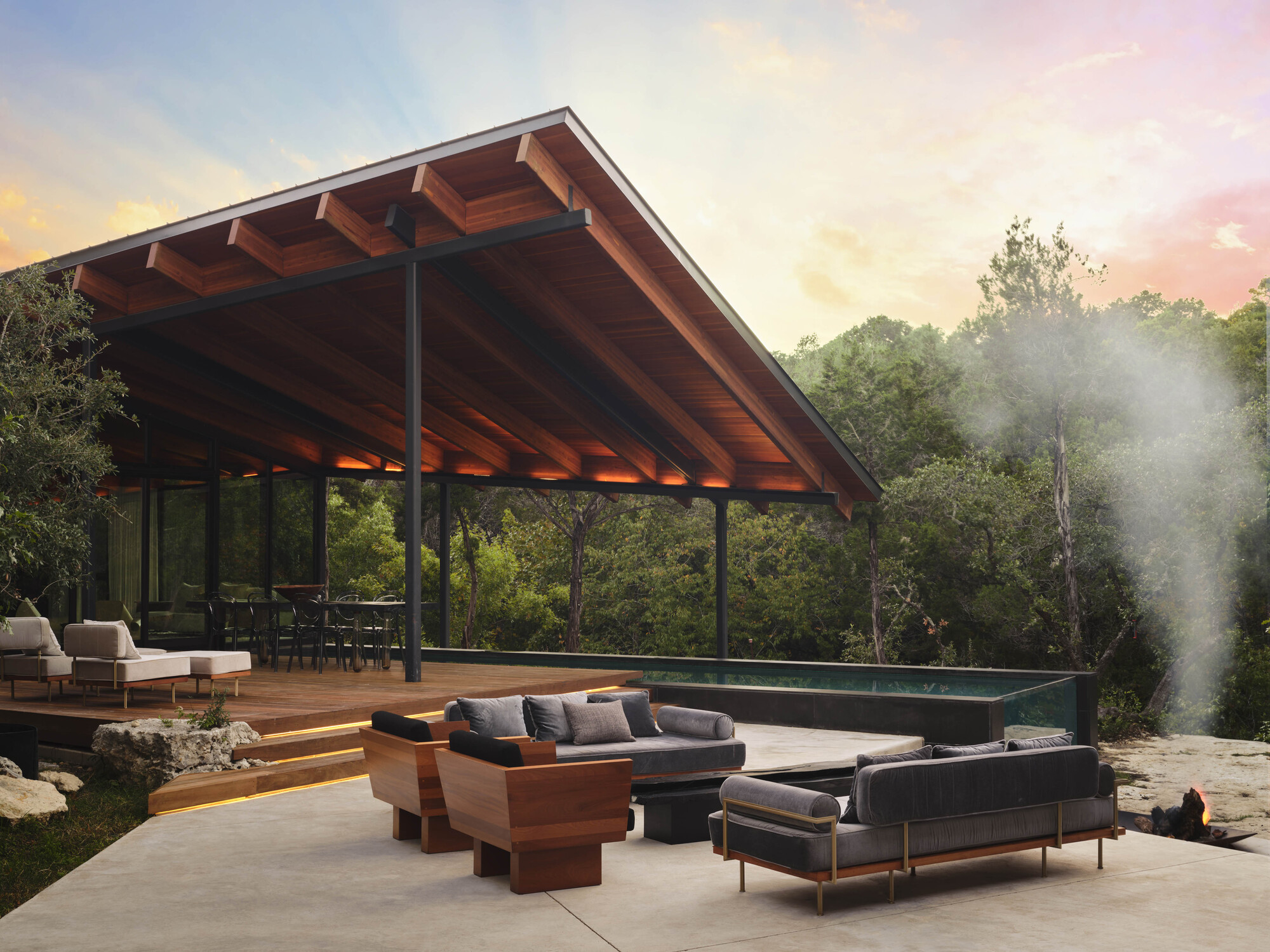 Open pavilion with exposed timber beams and steel columns overlooking a terrace with pool at sunset