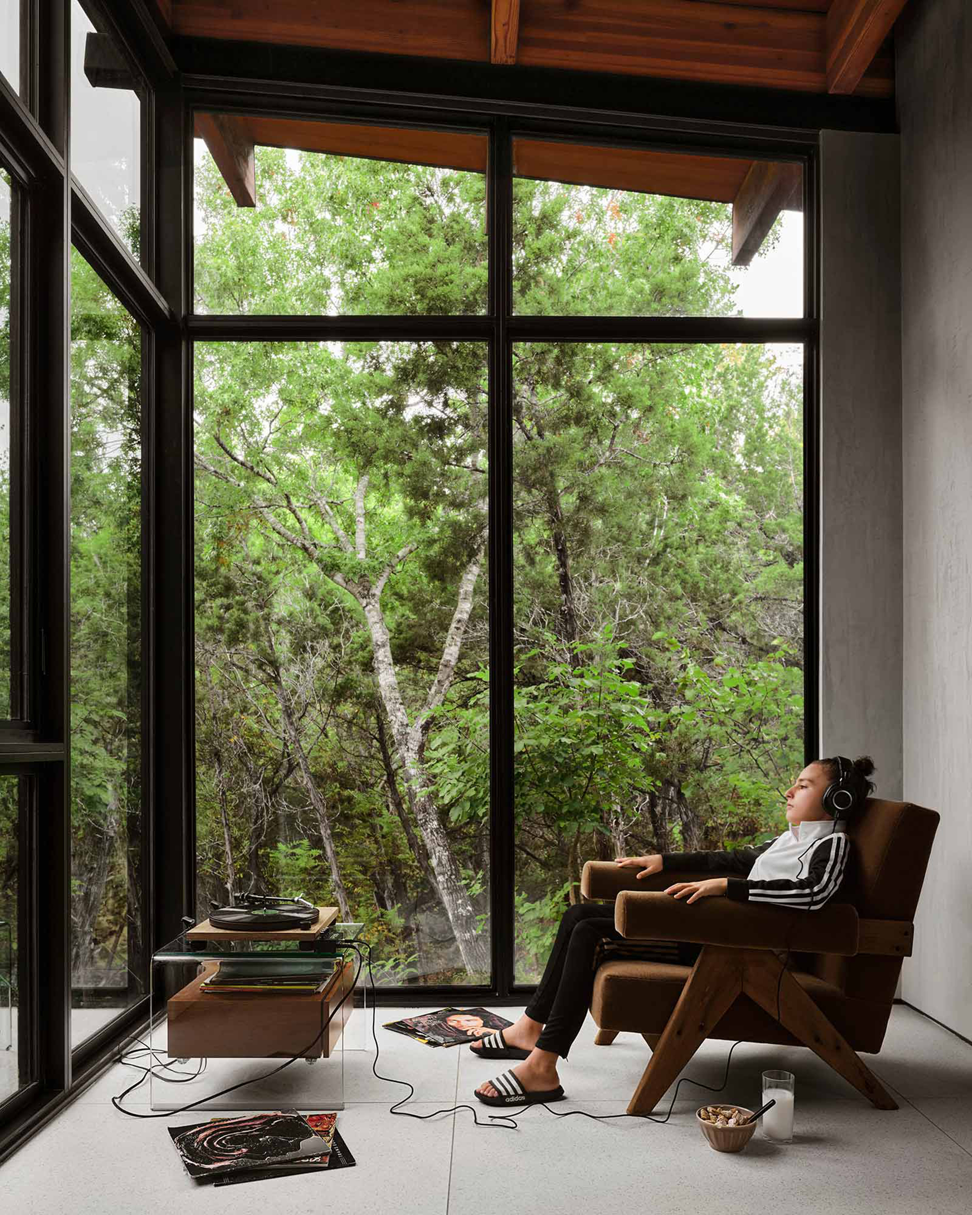 Interior seating nook with floor-to-ceiling windows framing trees where a person reclines listening to music