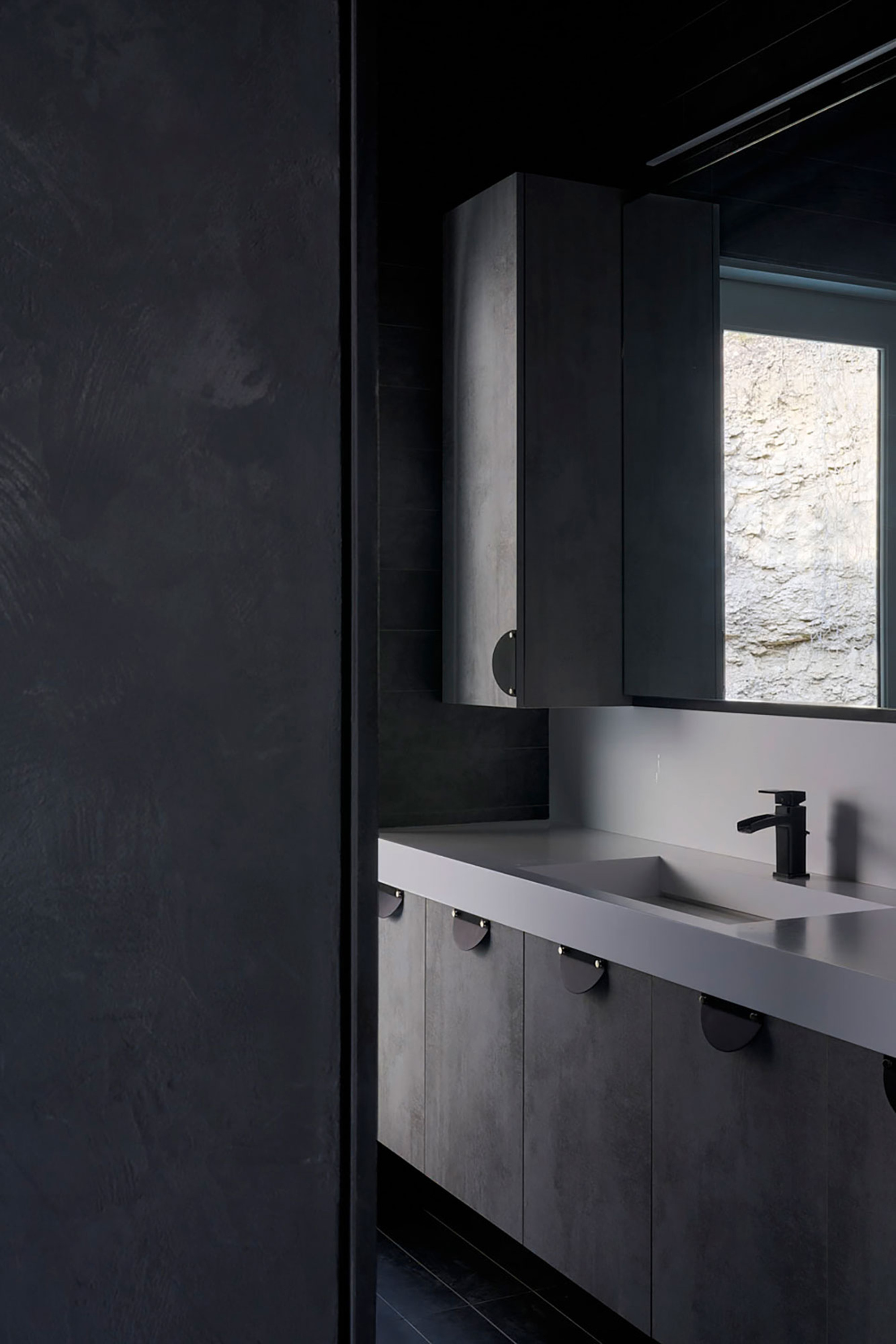 Bathroom vanity with concrete counter and narrow window revealing rough stone exterior wall