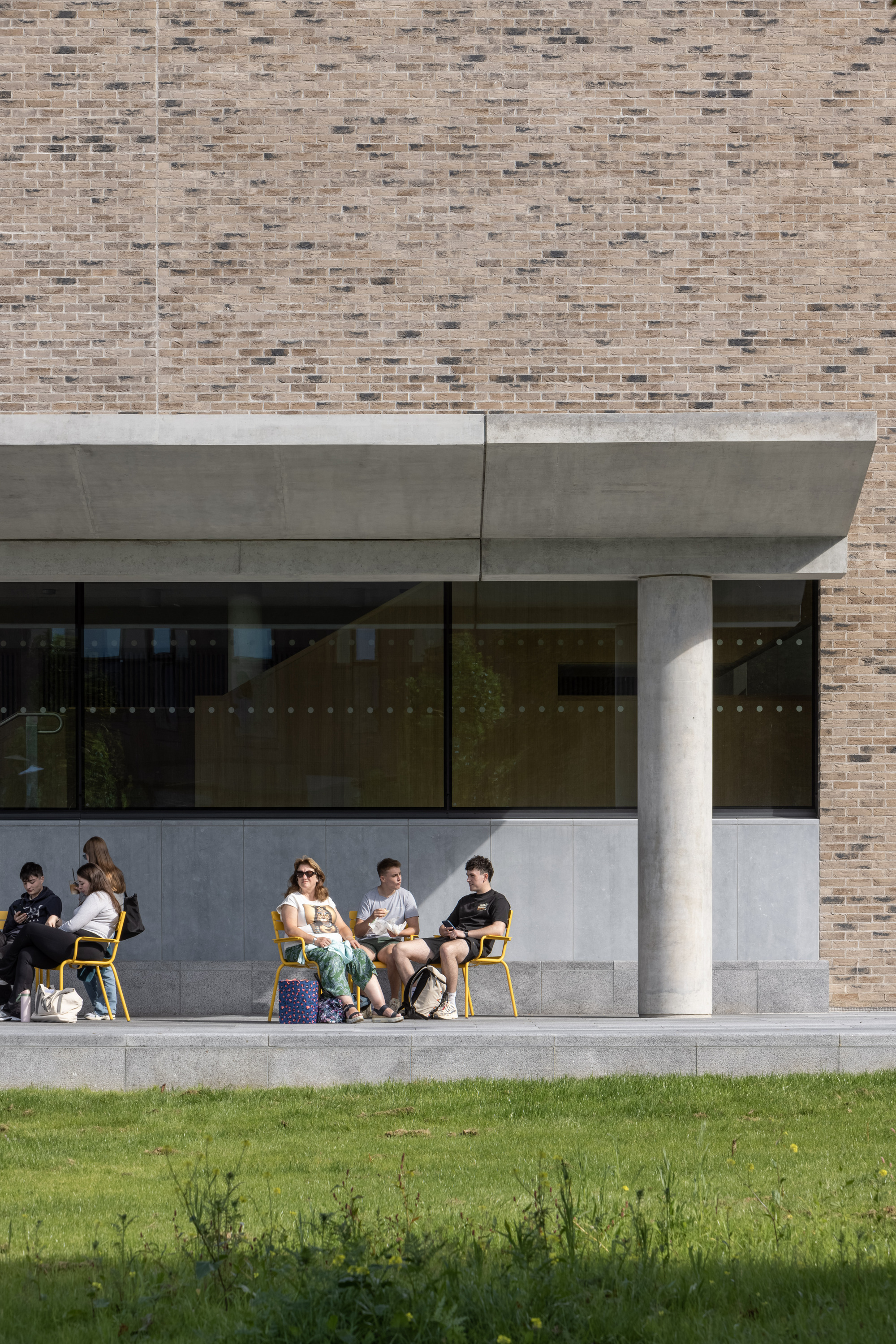 Covered terrace with concrete columns and beam supporting brick wall above students seated on yellow chairs