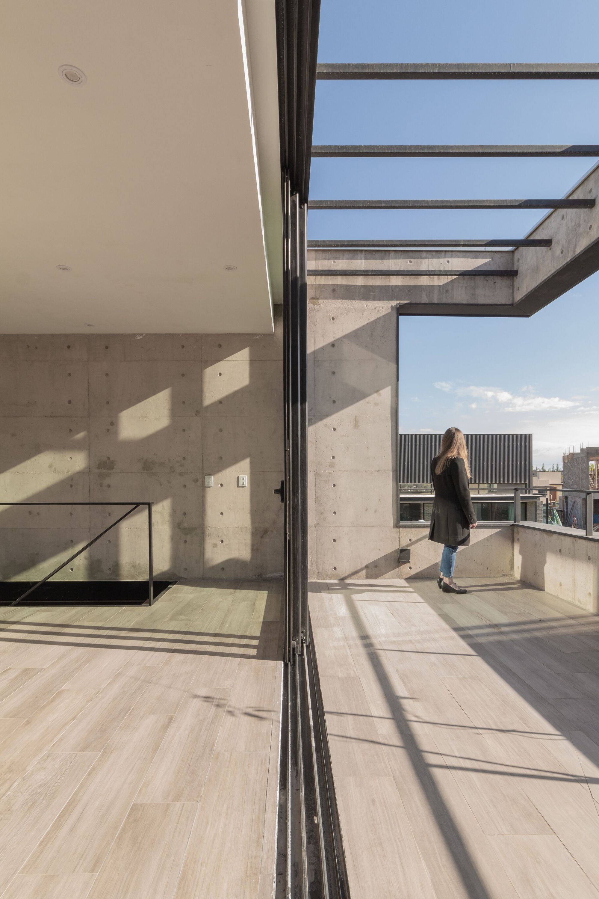 Sliding glass door opening to a concrete rooftop terrace with steel beam pergola and a person standing outside