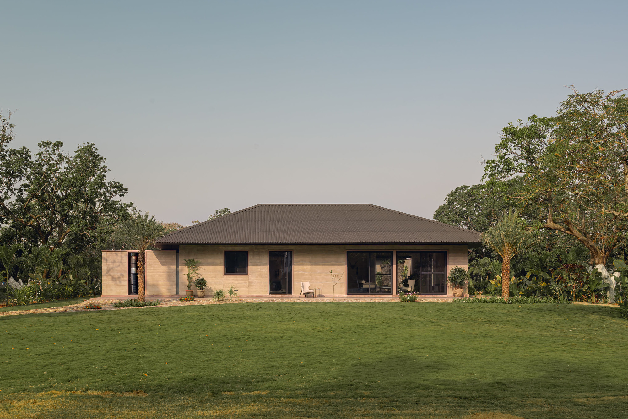 Rammed earth facade with hipped metal roof set against a manicured lawn and tree canopy at dusk