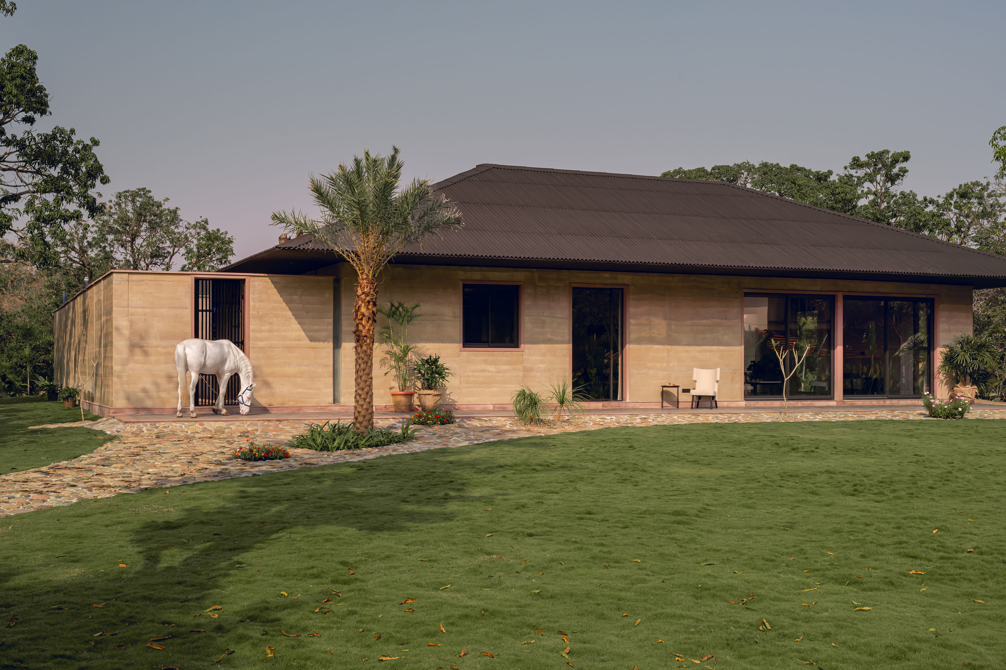 Garden facade with a white horse grazing beside palm trees and rammed earth walls at twilight