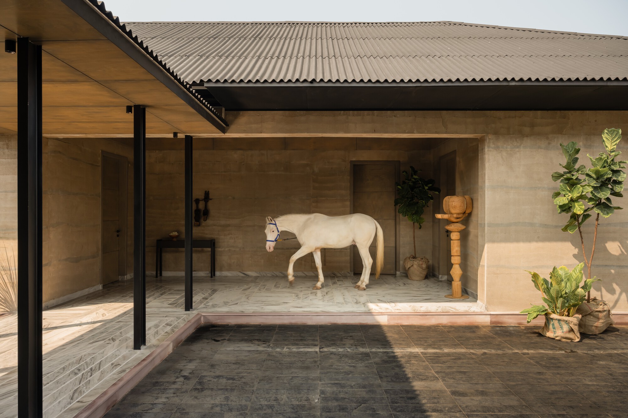 Covered courtyard with black steel columns and rammed earth walls sheltering a white horse in afternoon light