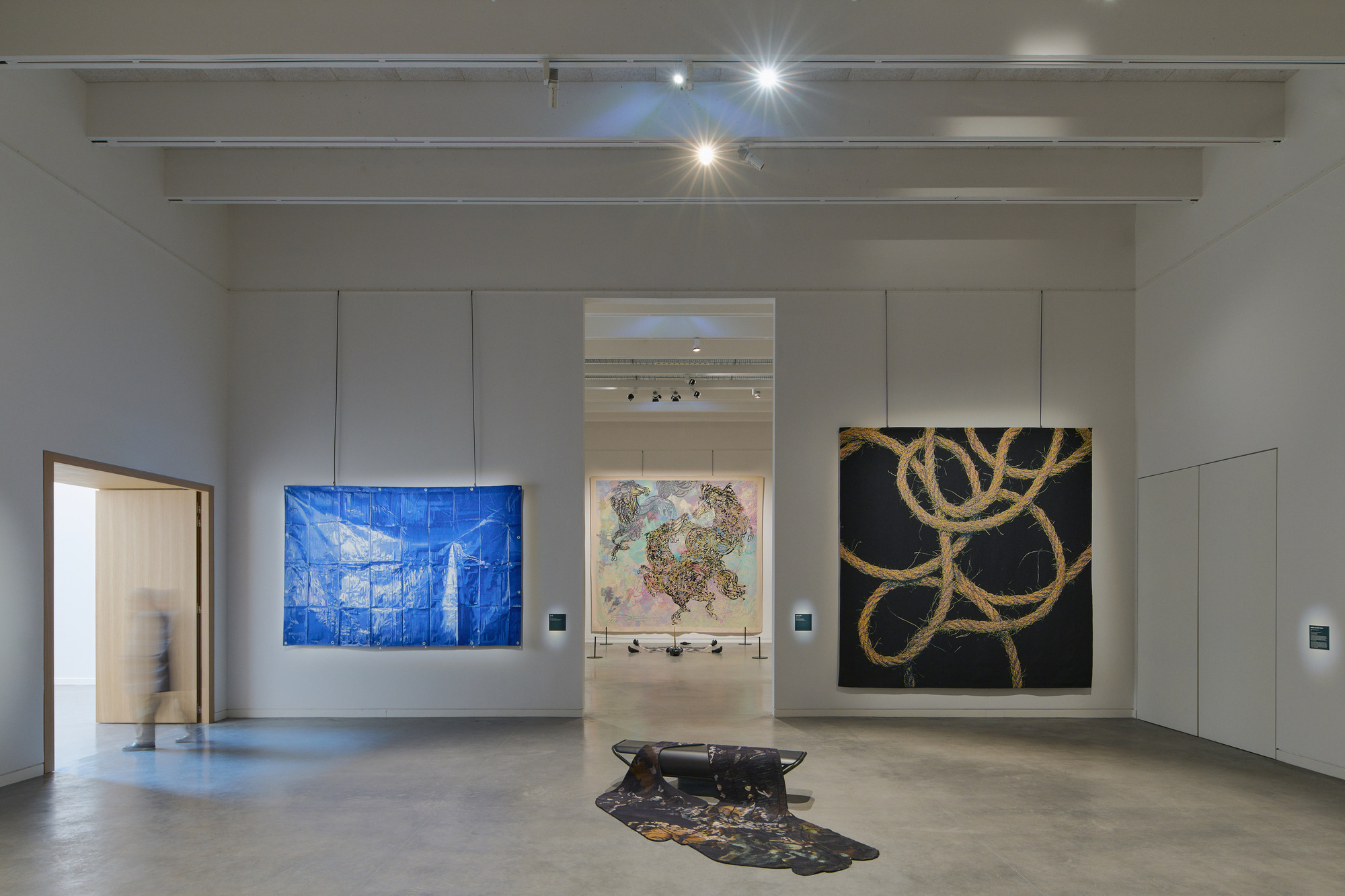 Gallery space with three large artworks on white walls and track lighting above polished concrete floors