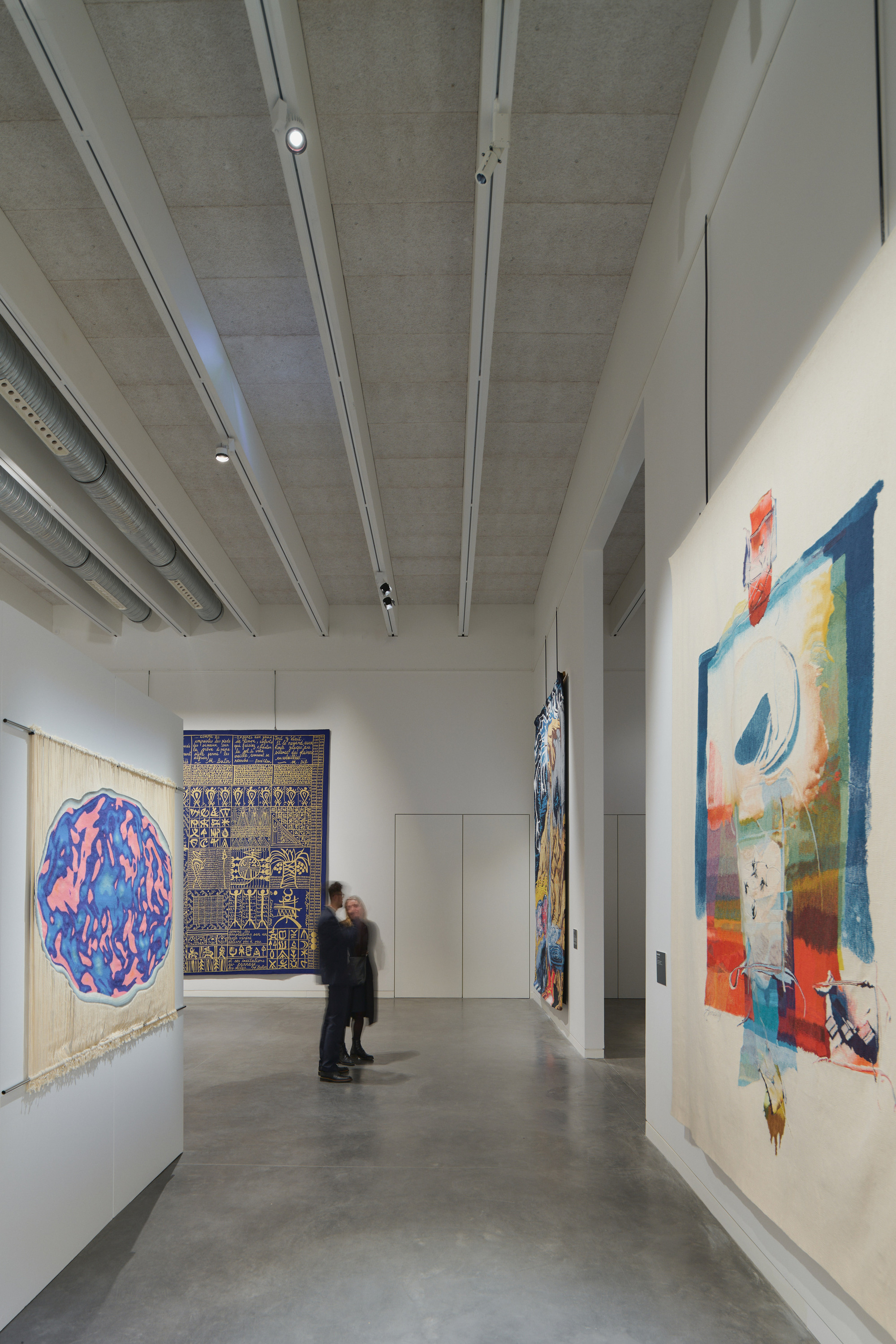 Gallery corridor with exposed concrete ceiling, white beams, and polished concrete floors displaying textile artworks