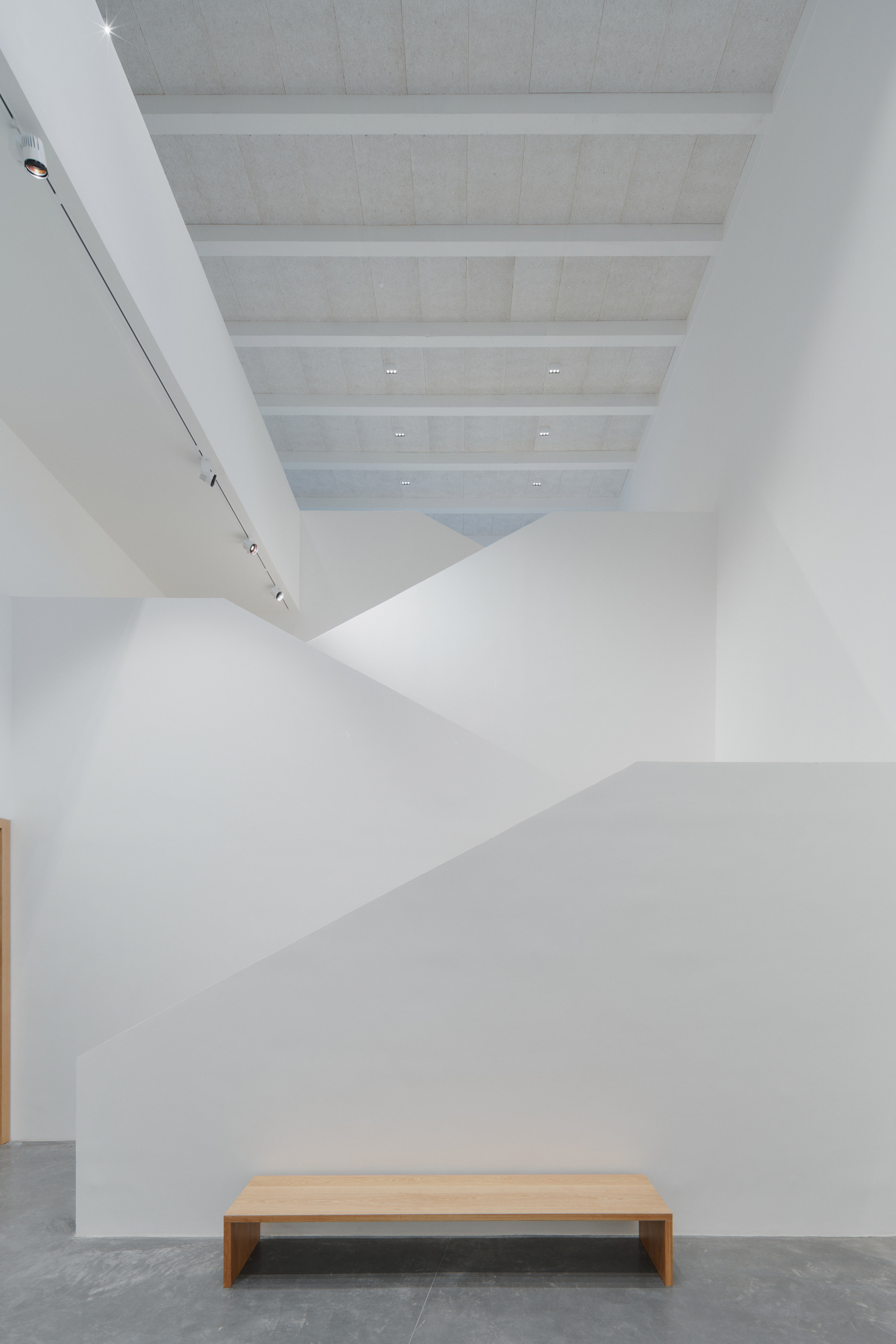 White gallery wall with angled ceiling planes meeting above a timber bench on concrete floor