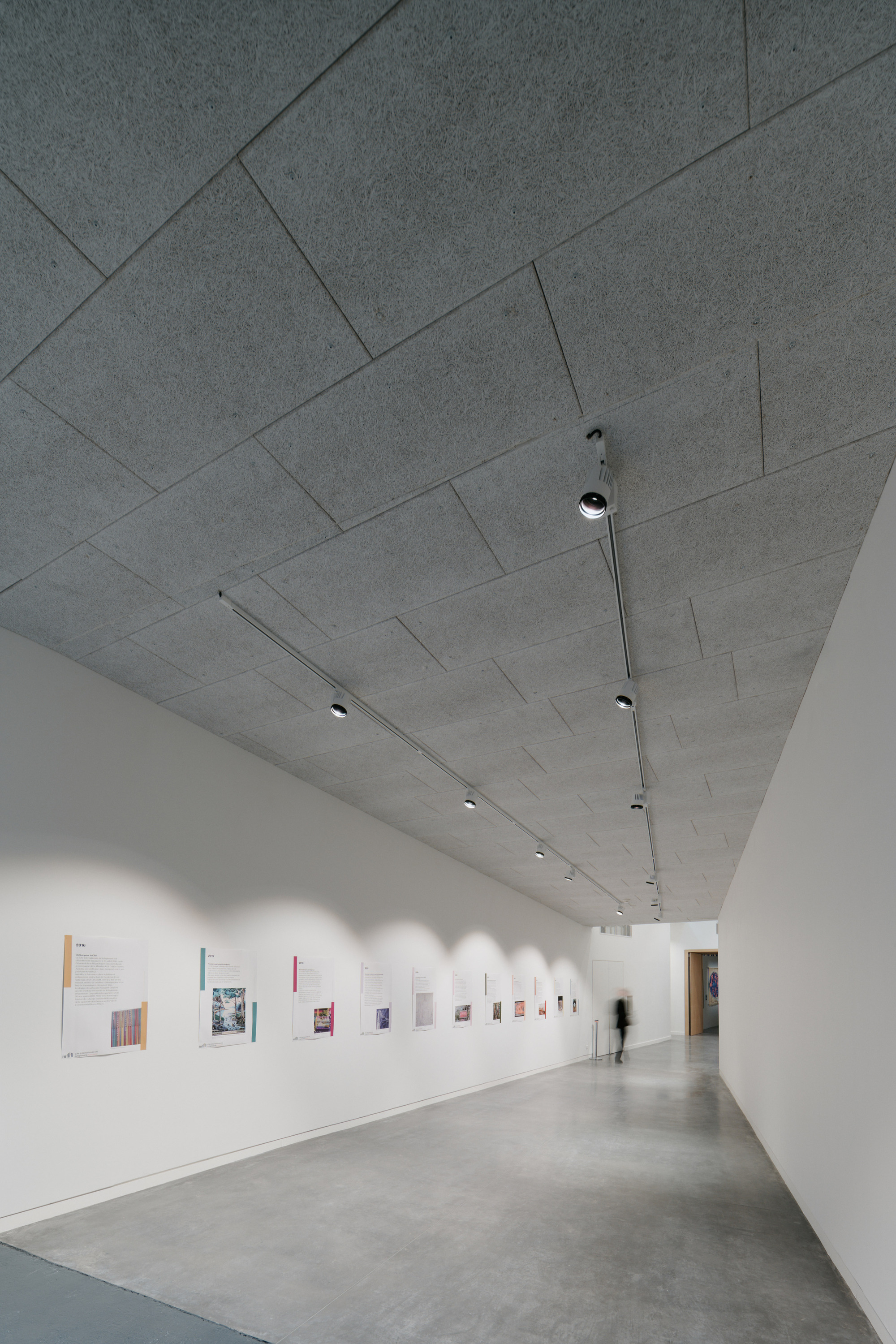 Gallery hallway with rectangular concrete ceiling panels and a figure passing small framed works on white walls