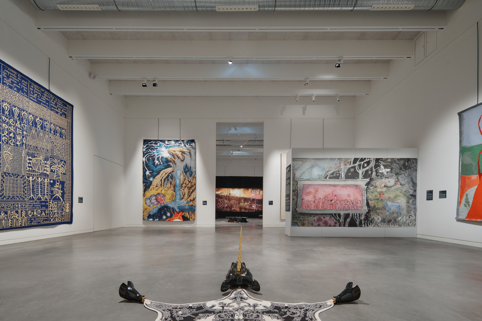 Exhibition hall with white ceiling beams and floor sculpture beneath hanging artworks arranged around doorways