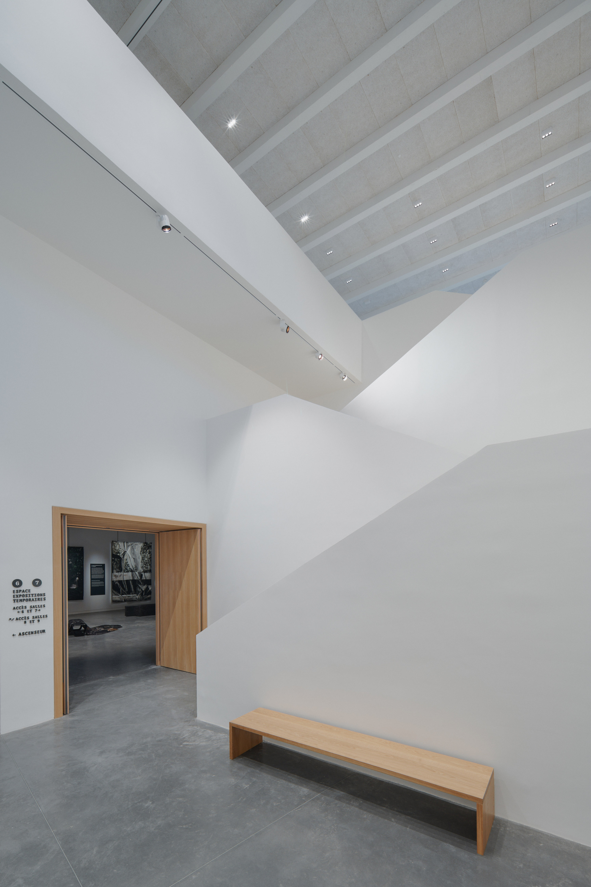 Gallery entrance with timber doorway and bench beneath a ribbed skylight and white walls