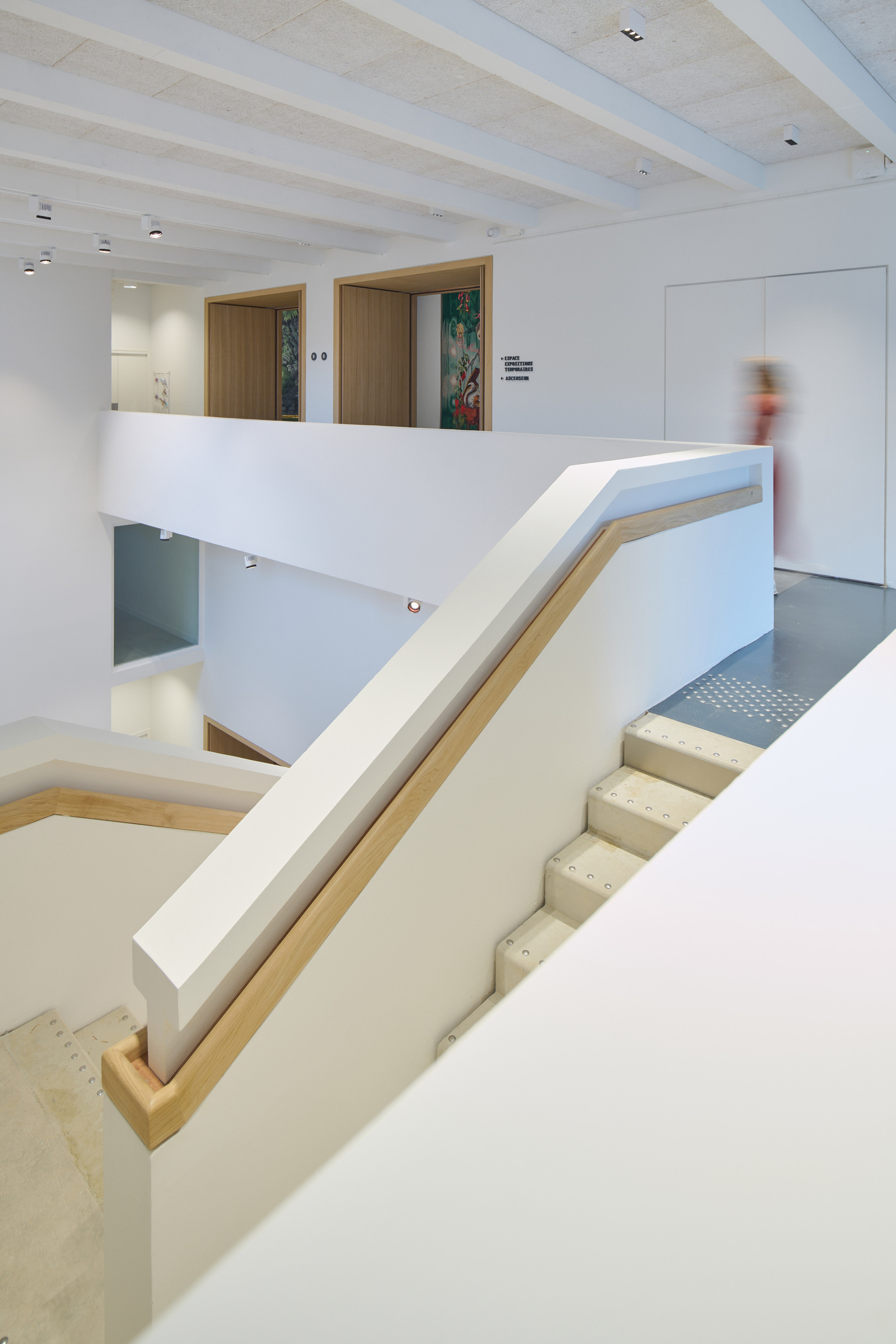 Staircase with white walls and timber handrail connecting multiple levels with a blurred visitor