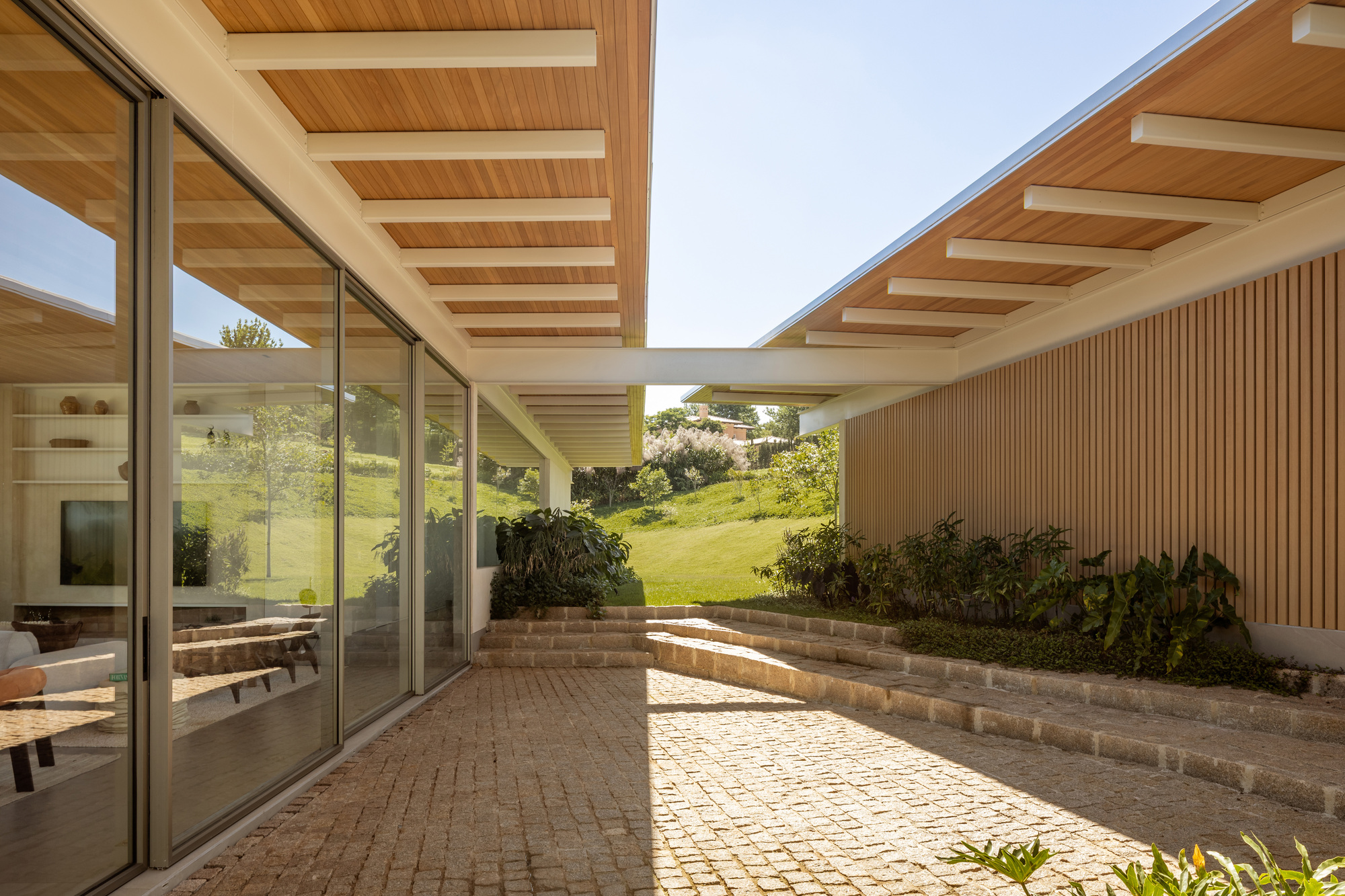 Brick-paved courtyard framed by timber slatted walls and floor-to-ceiling glass under exposed beam ceilings