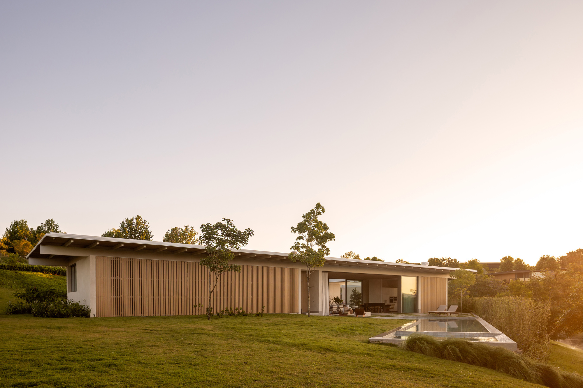 Long low-slung volume with horizontal wood cladding and projecting roof set into a sloping lawn at sunset