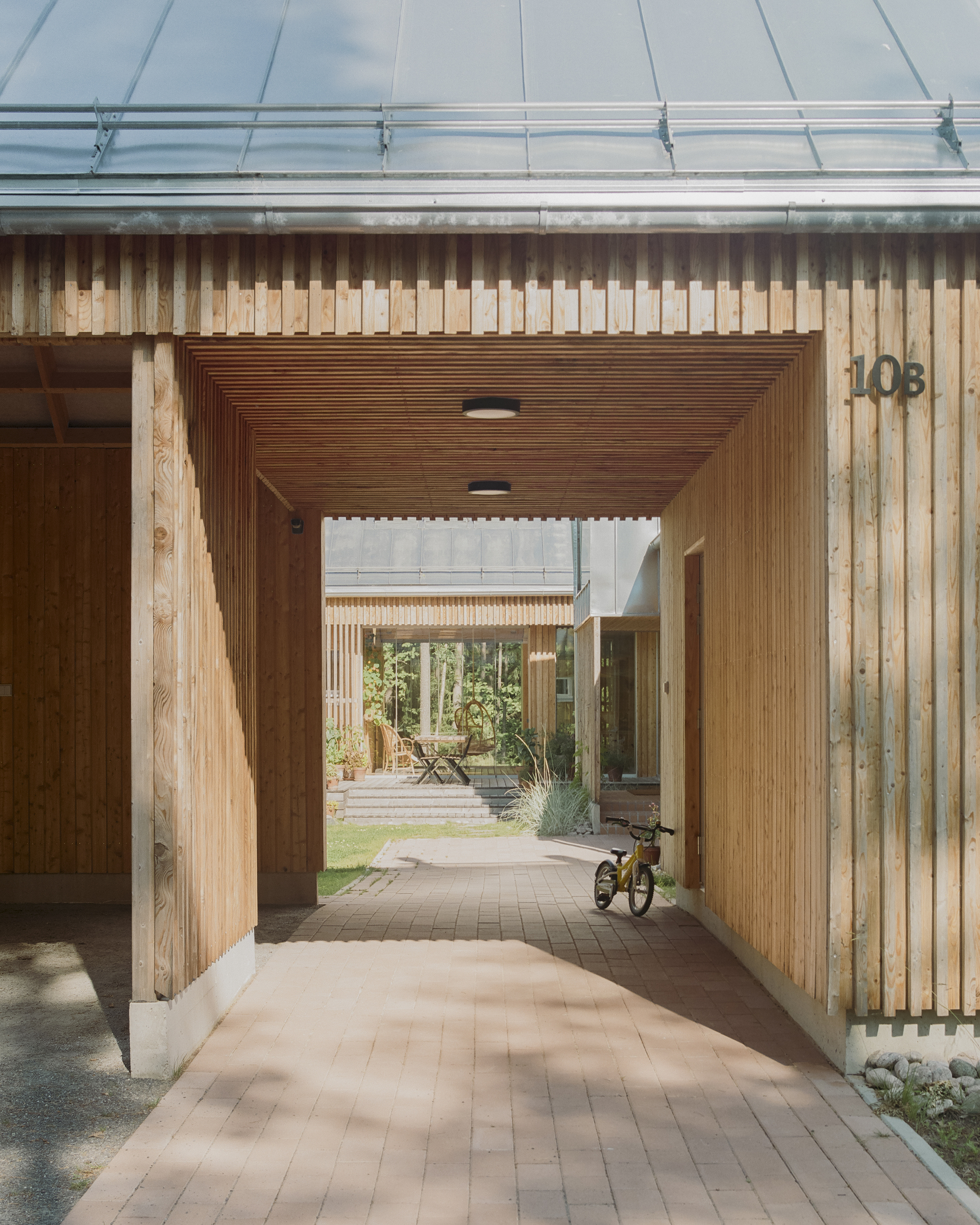 Covered timber passageway with vertical slatted walls leading to an interior courtyard with a tricycle