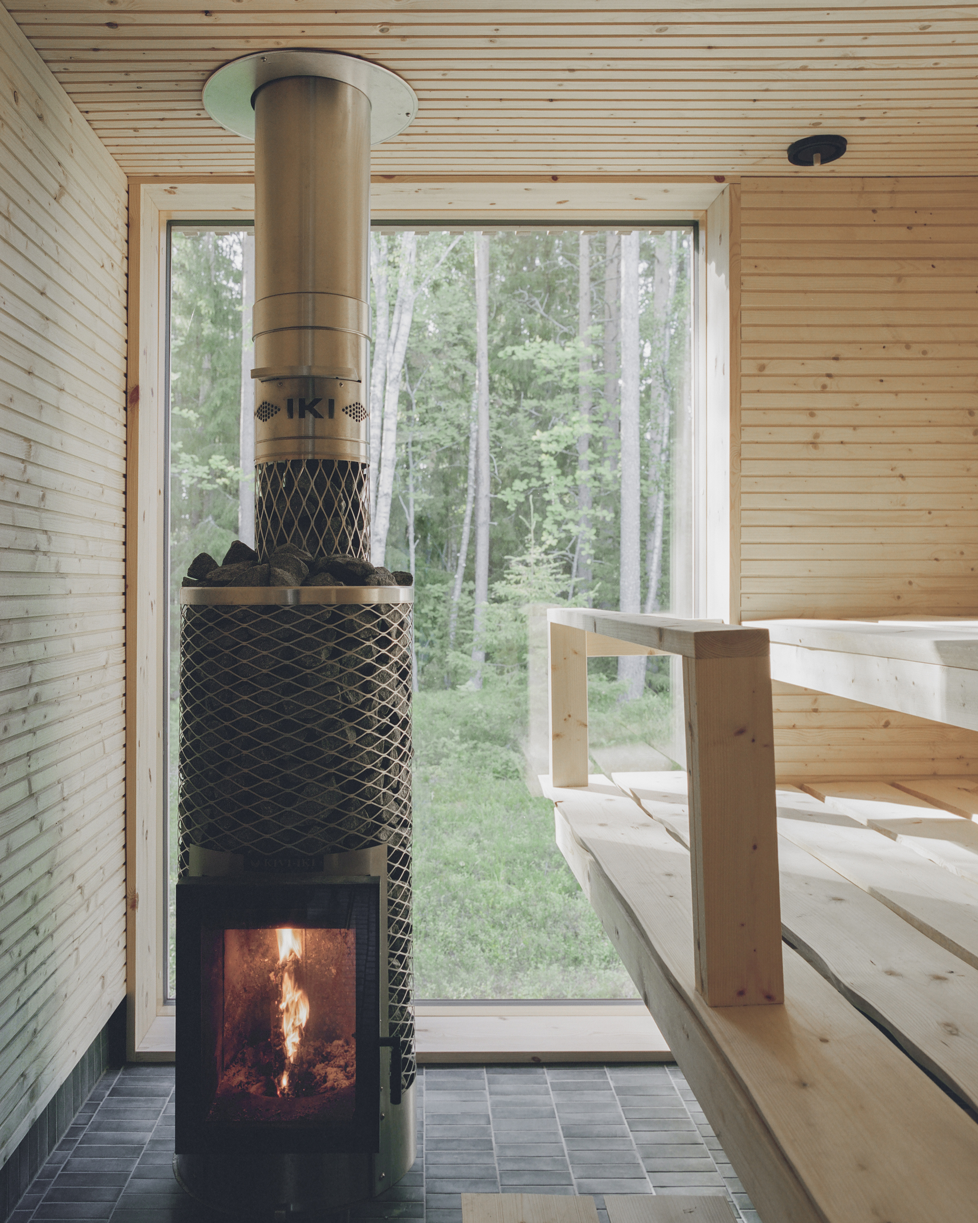 Mesh-wrapped wood-burning sauna stove with glass door showing flames, adjacent to tiered timber benches overlooking forest