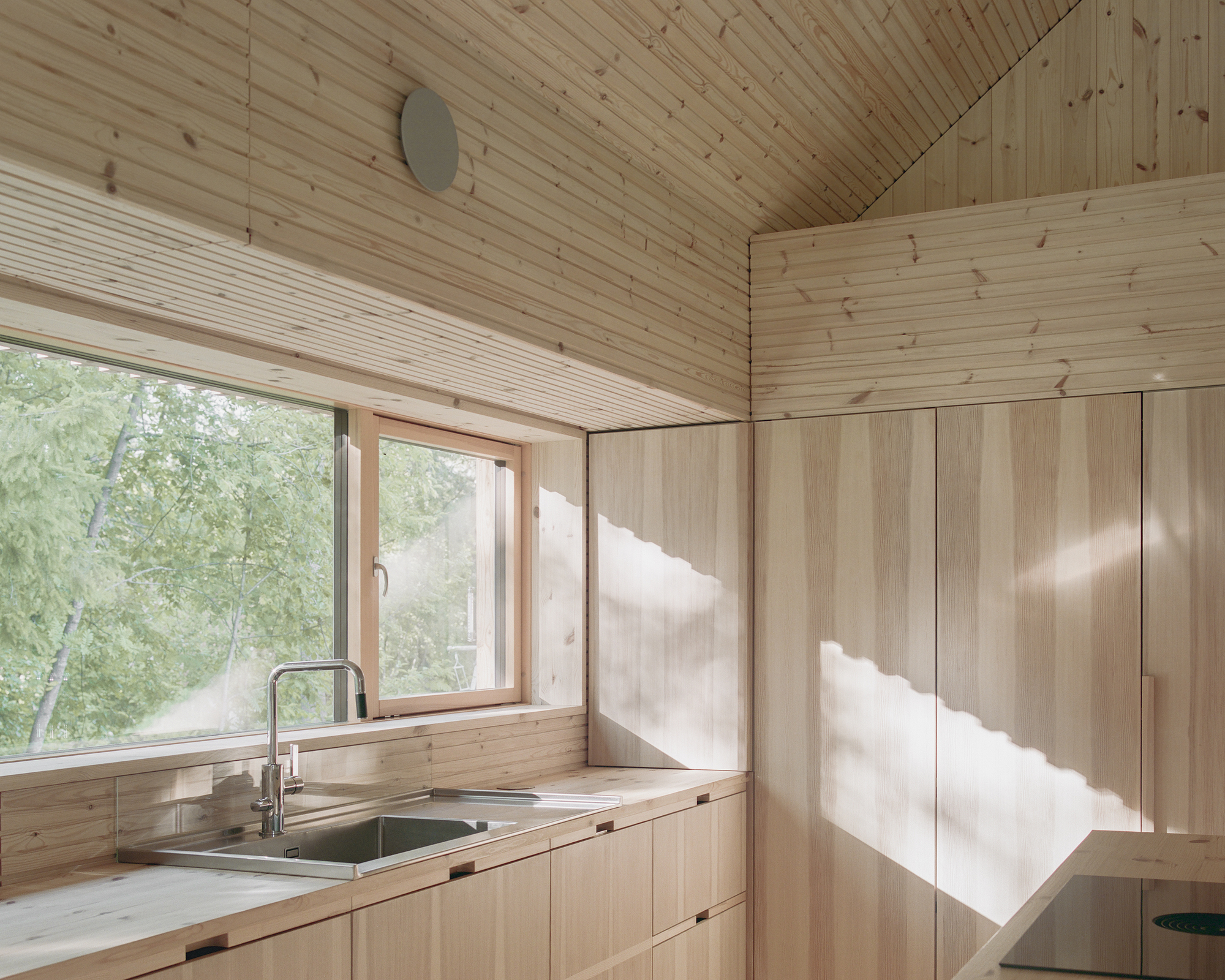 Kitchen corner with timber countertop and sink under window, sunlight casting shadows across plywood cabinetry