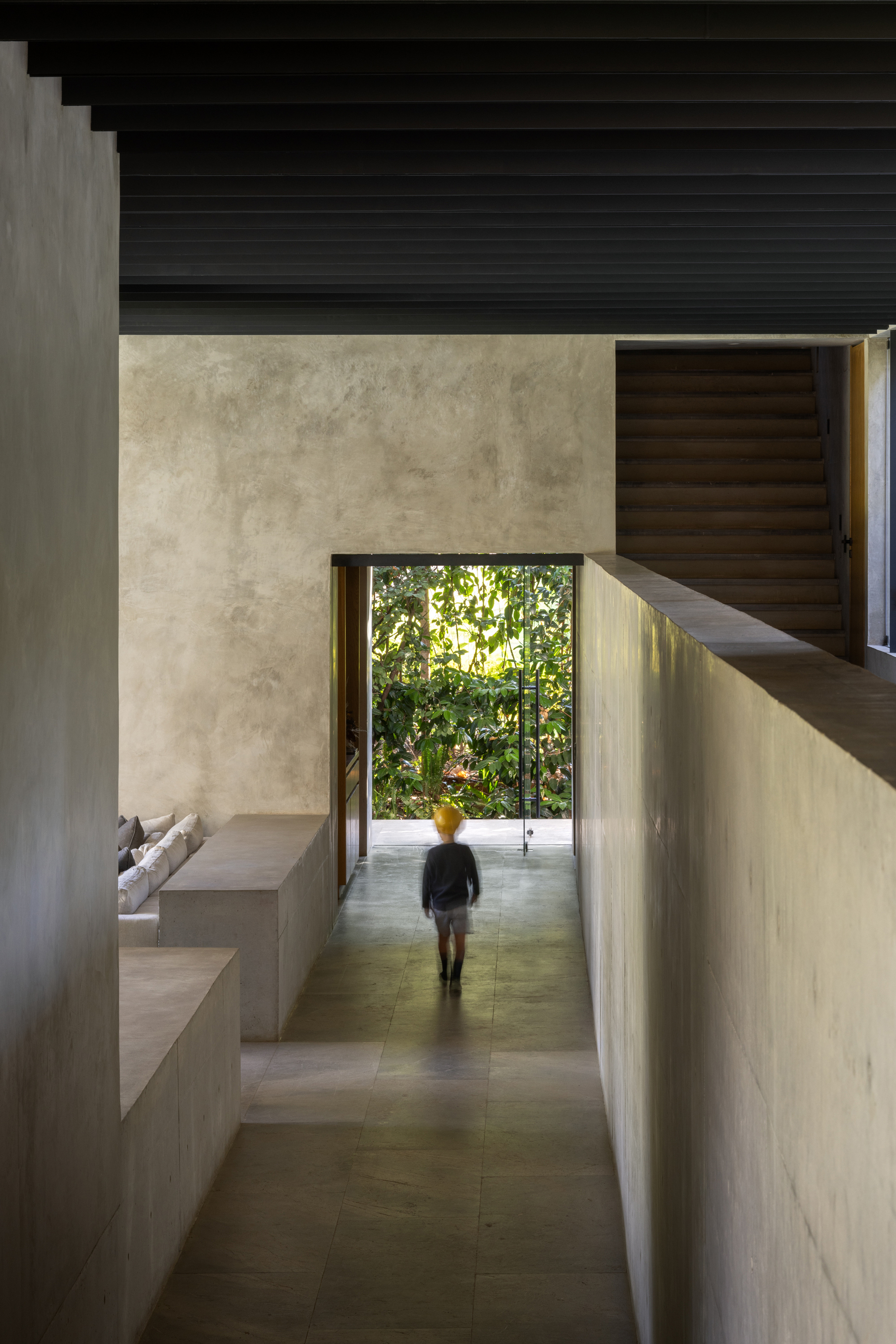 Entry corridor with figure walking toward garden framed by concrete walls and dark timber ceiling