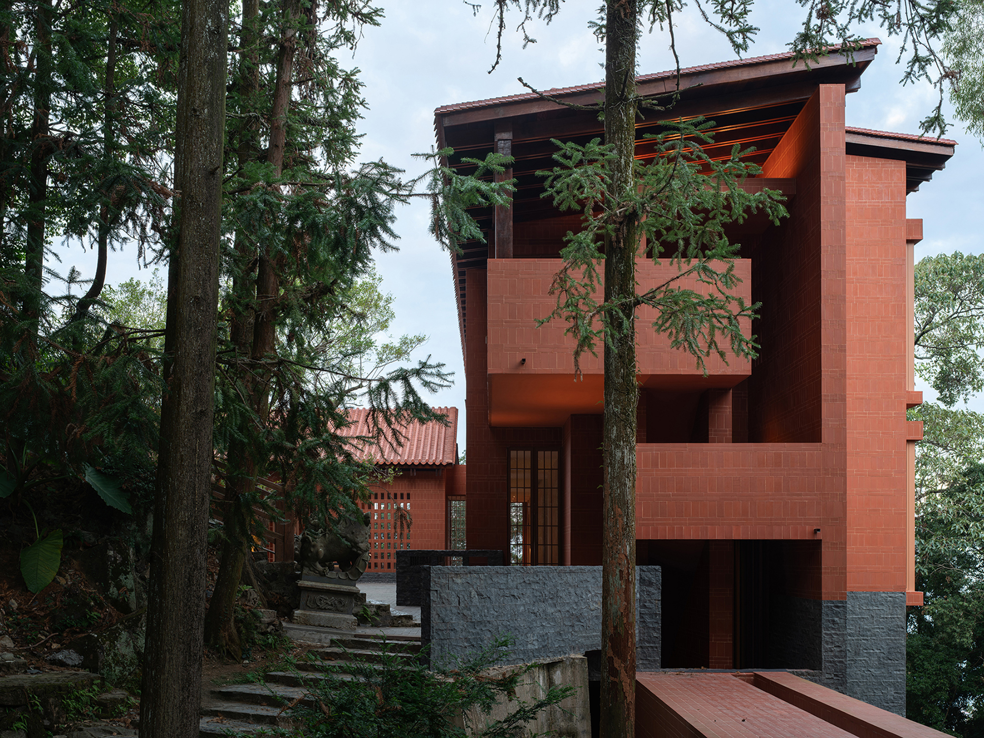 Multi-level red brick volume with exposed timber roof framing rising through tall pine trees
