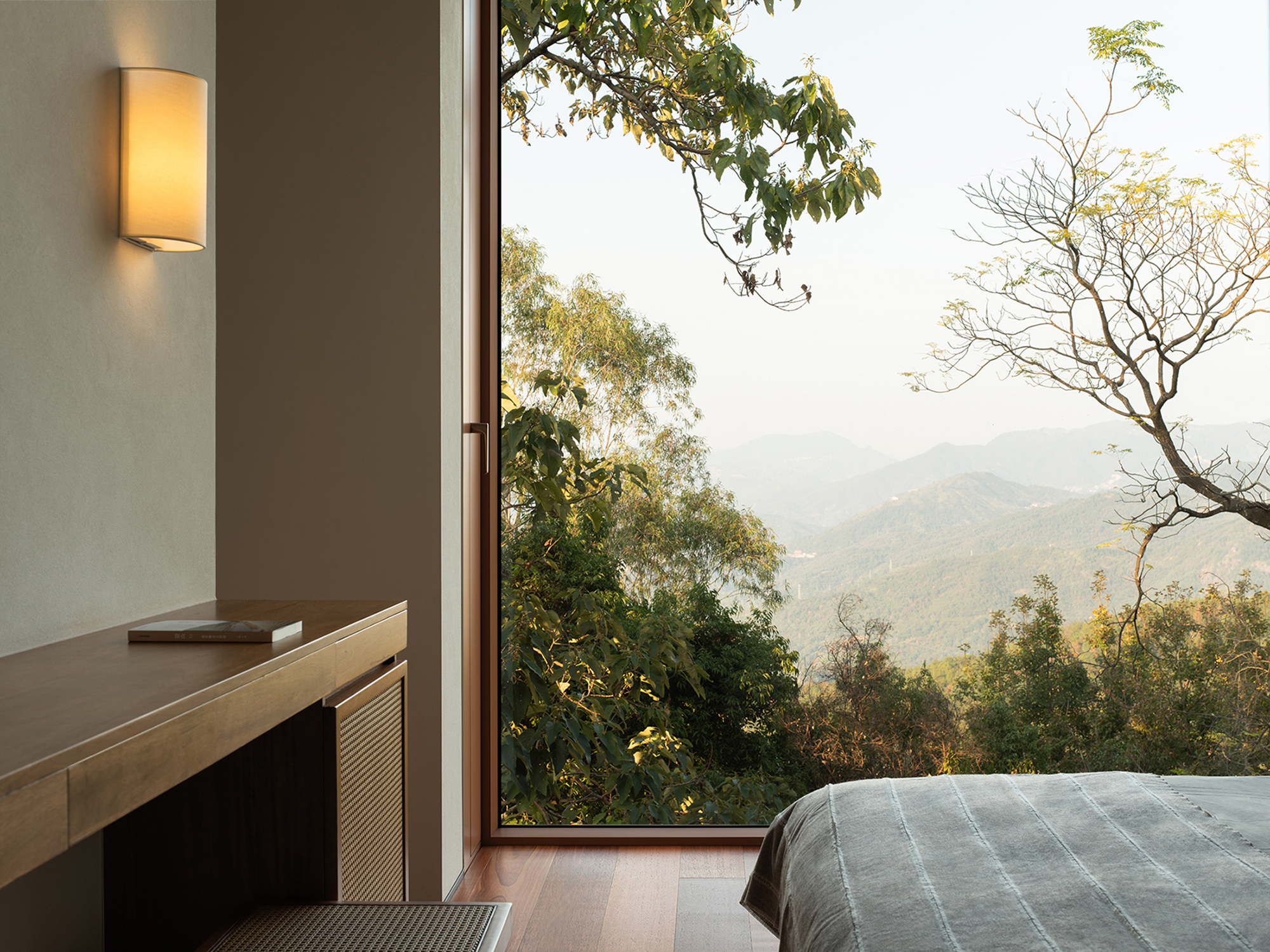 Floor-to-ceiling bedroom window overlooking forested hillside and distant mountains in daylight