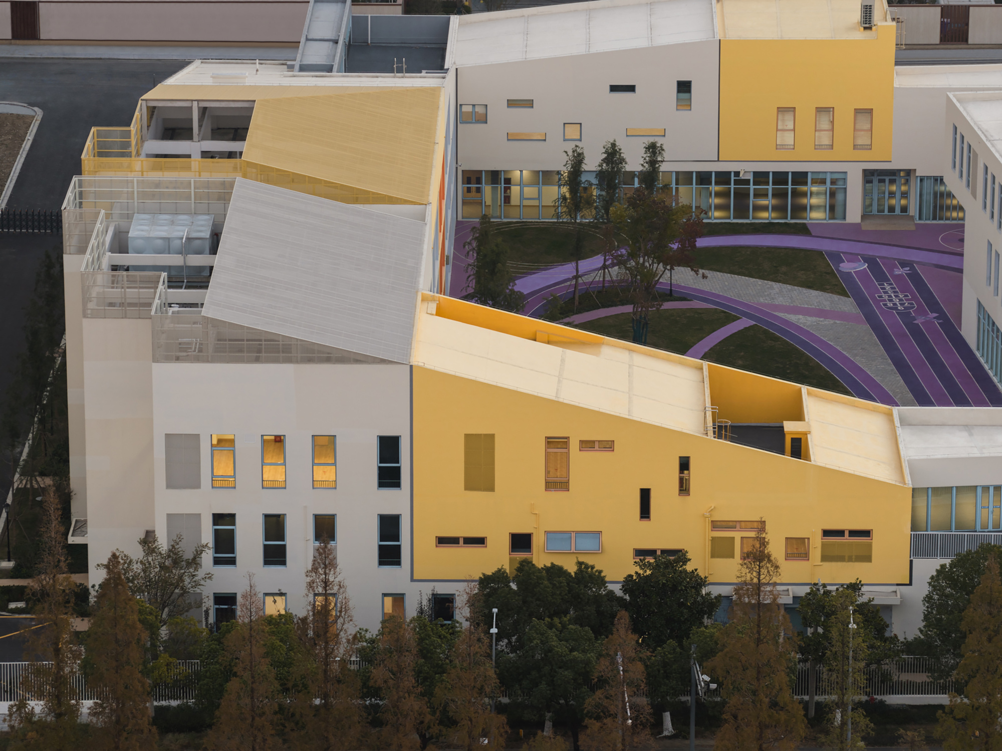Facade composition with yellow and white panels framing a planted courtyard with purple sports markings at dusk