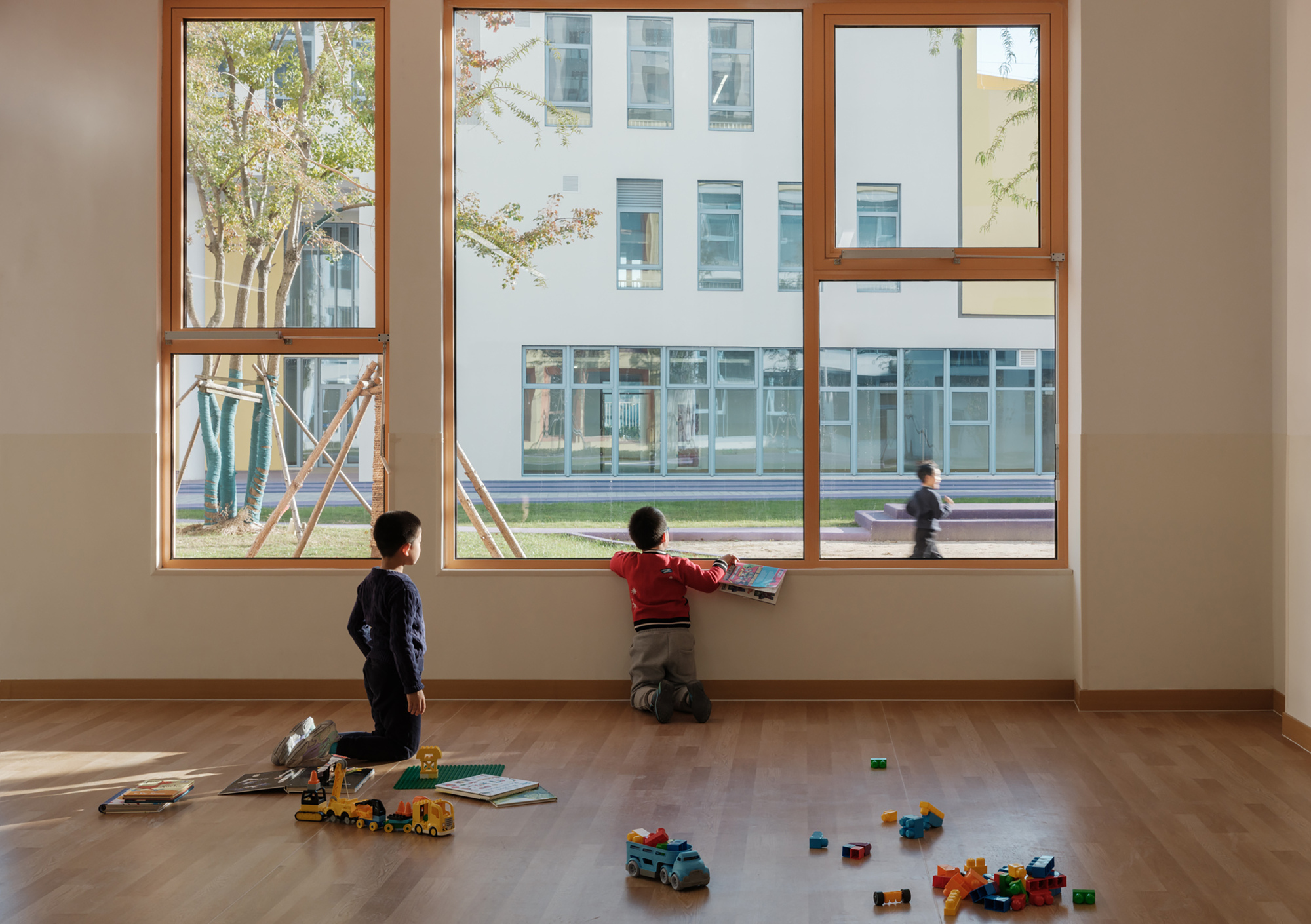 Interior classroom with timber-framed windows overlooking the courtyard where two children sit with scattered toys on the floor