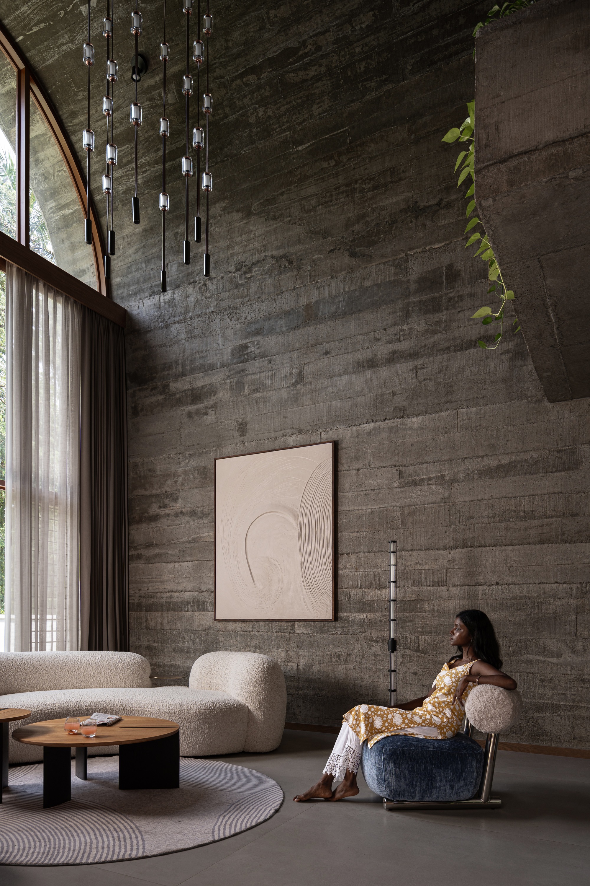Double-height living space with board-formed concrete wall, pendant light installation and person seated in armchair