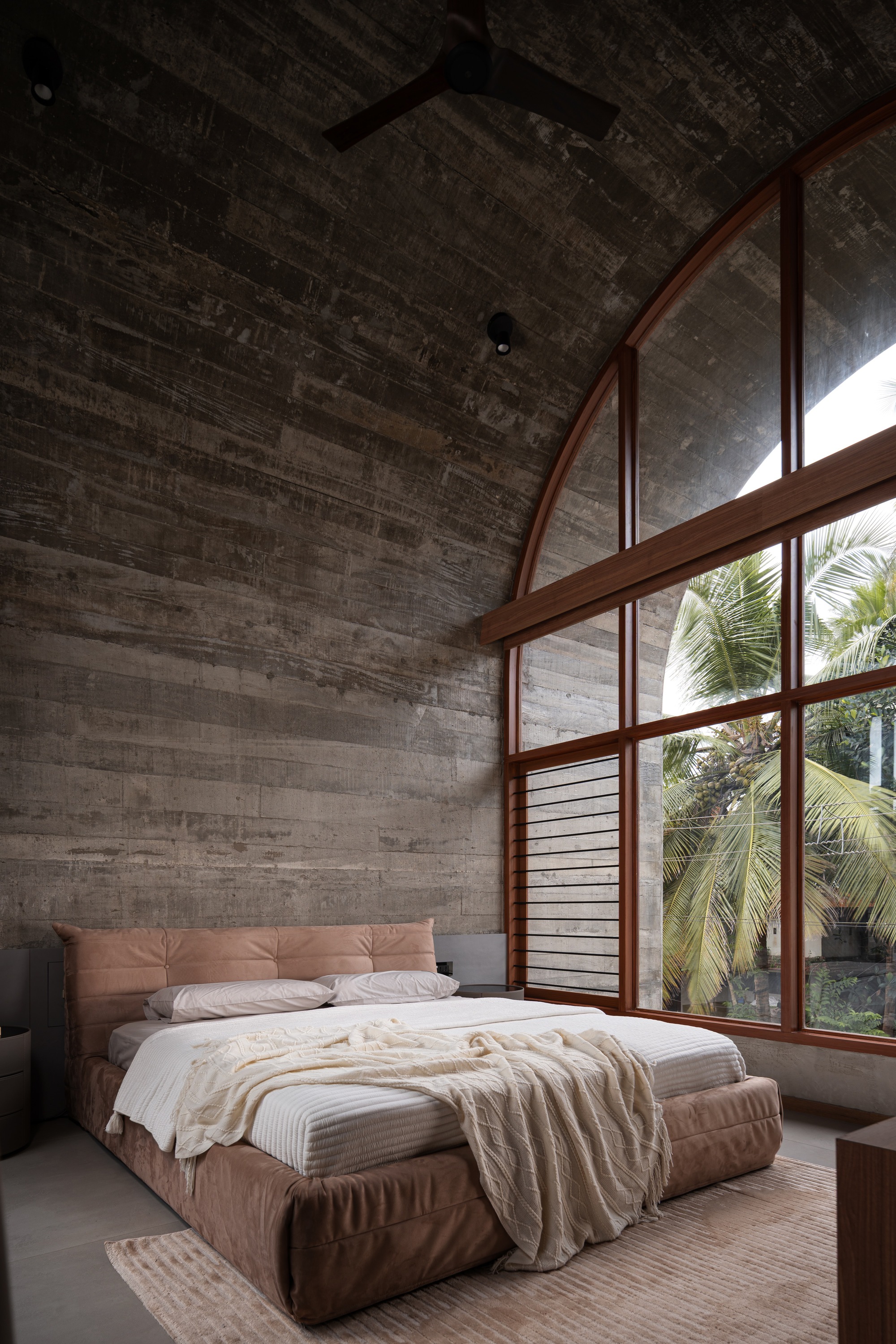 Bedroom with arched timber-framed windows overlooking palm trees beneath a board-formed concrete vaulted ceiling