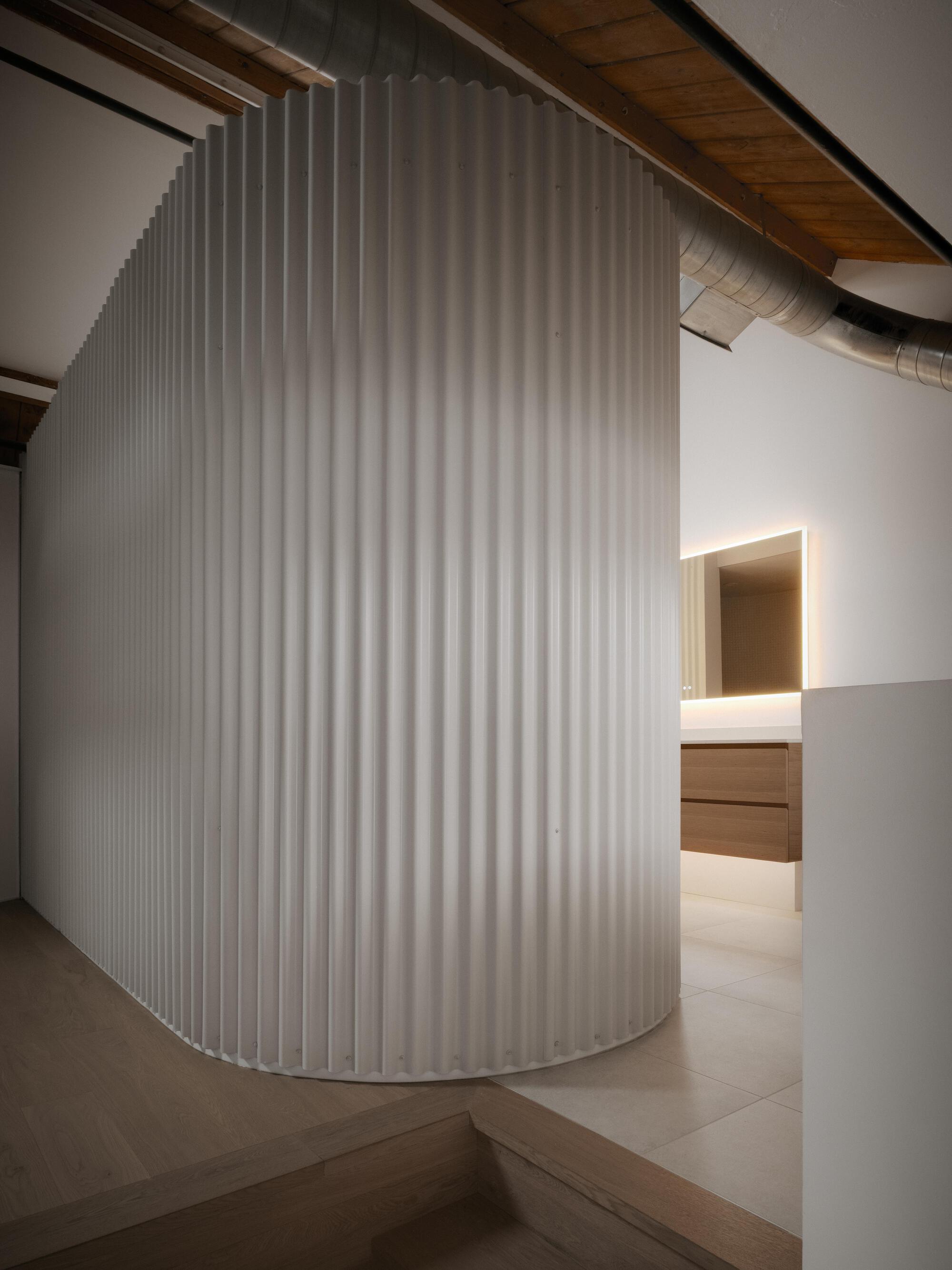 Curved white louver screen enclosing volume with glimpse of backlit timber cabinetry beyond