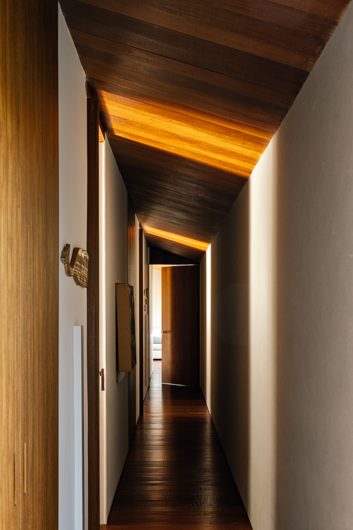 Corridor with folded timber ceiling planes and concealed lighting washing the white walls at dusk