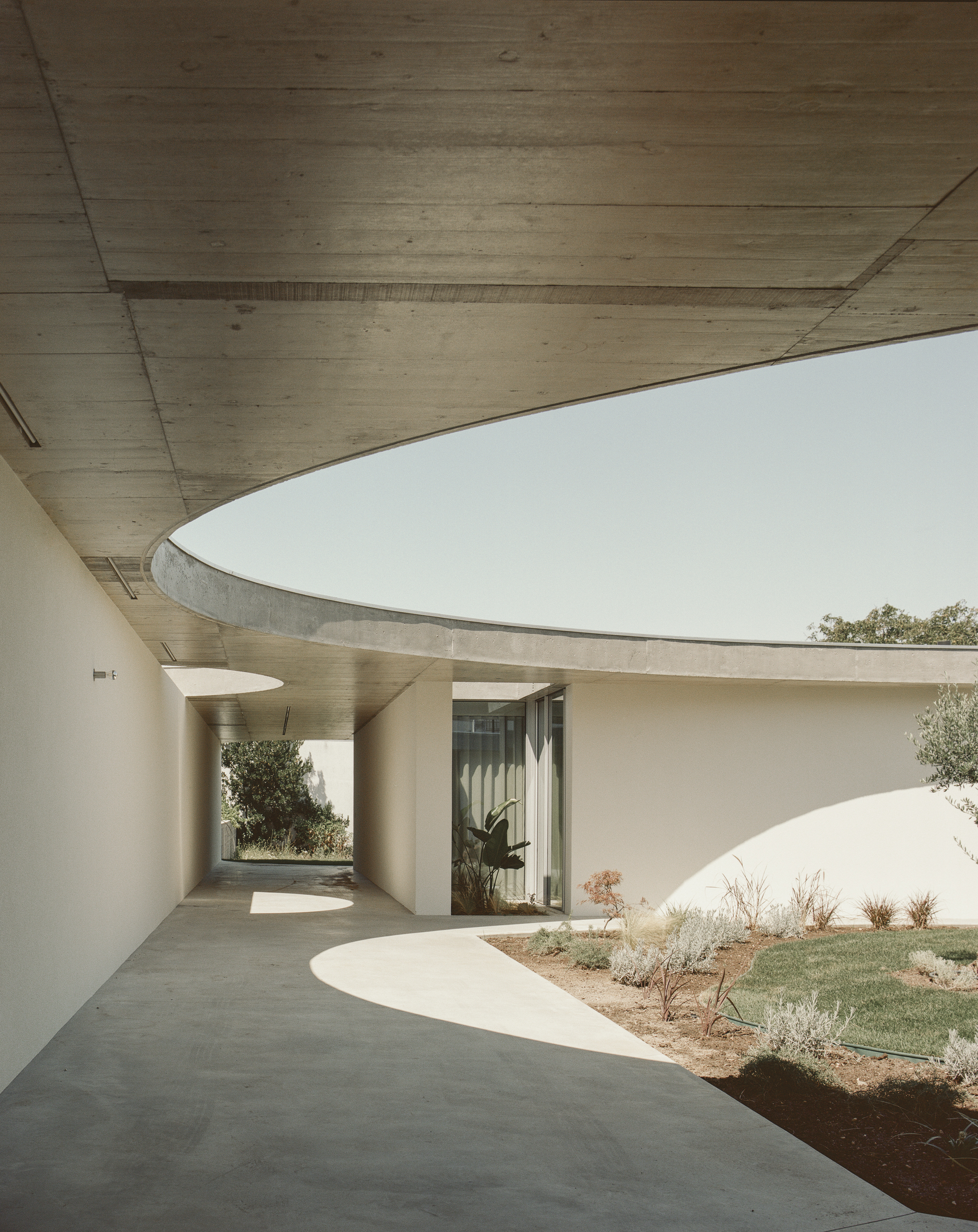 Curved covered walkway beneath exposed concrete ceiling with courtyard and plantings beyond