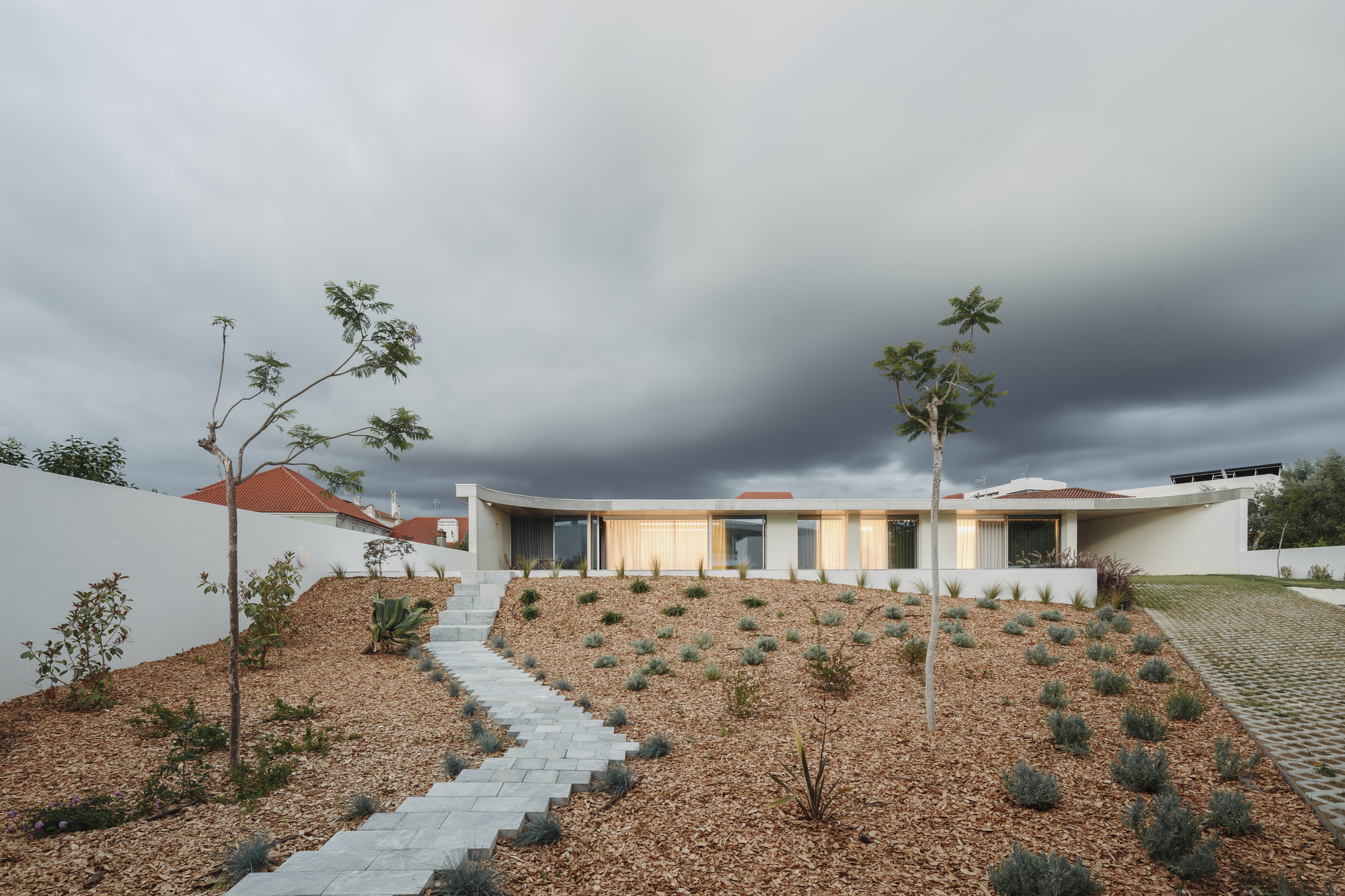 Concrete paving steps ascending through a mulched hillside planted with low shrubs and young trees under stormy skies