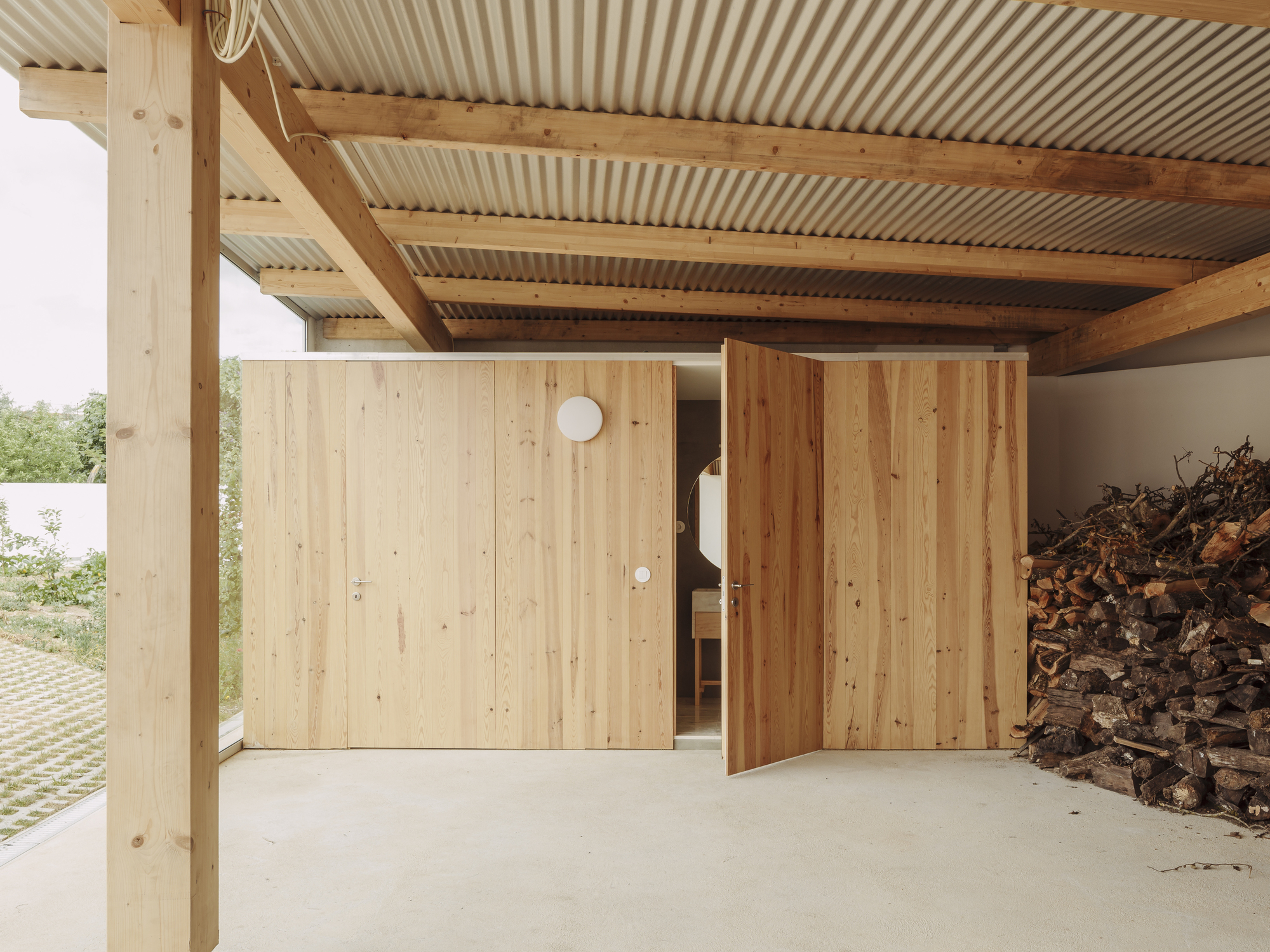 Covered carport with timber posts, exposed beams, corrugated metal ceiling, and sliding wood panel doors beside stacked firewood