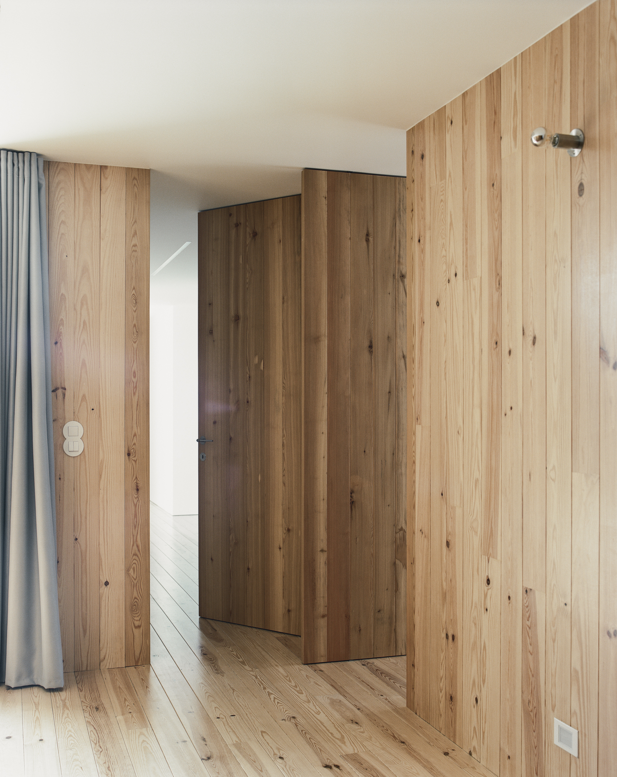 Vertical timber wall panels concealing a door beside a grey curtain under soft daylight