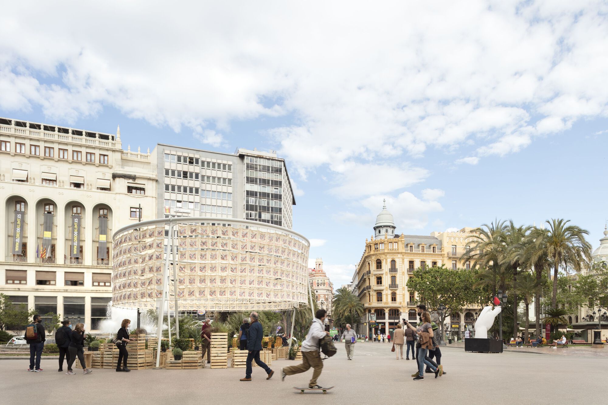 Cylindrical fabric pavilion with printed textile panels in a public plaza surrounded by palm trees