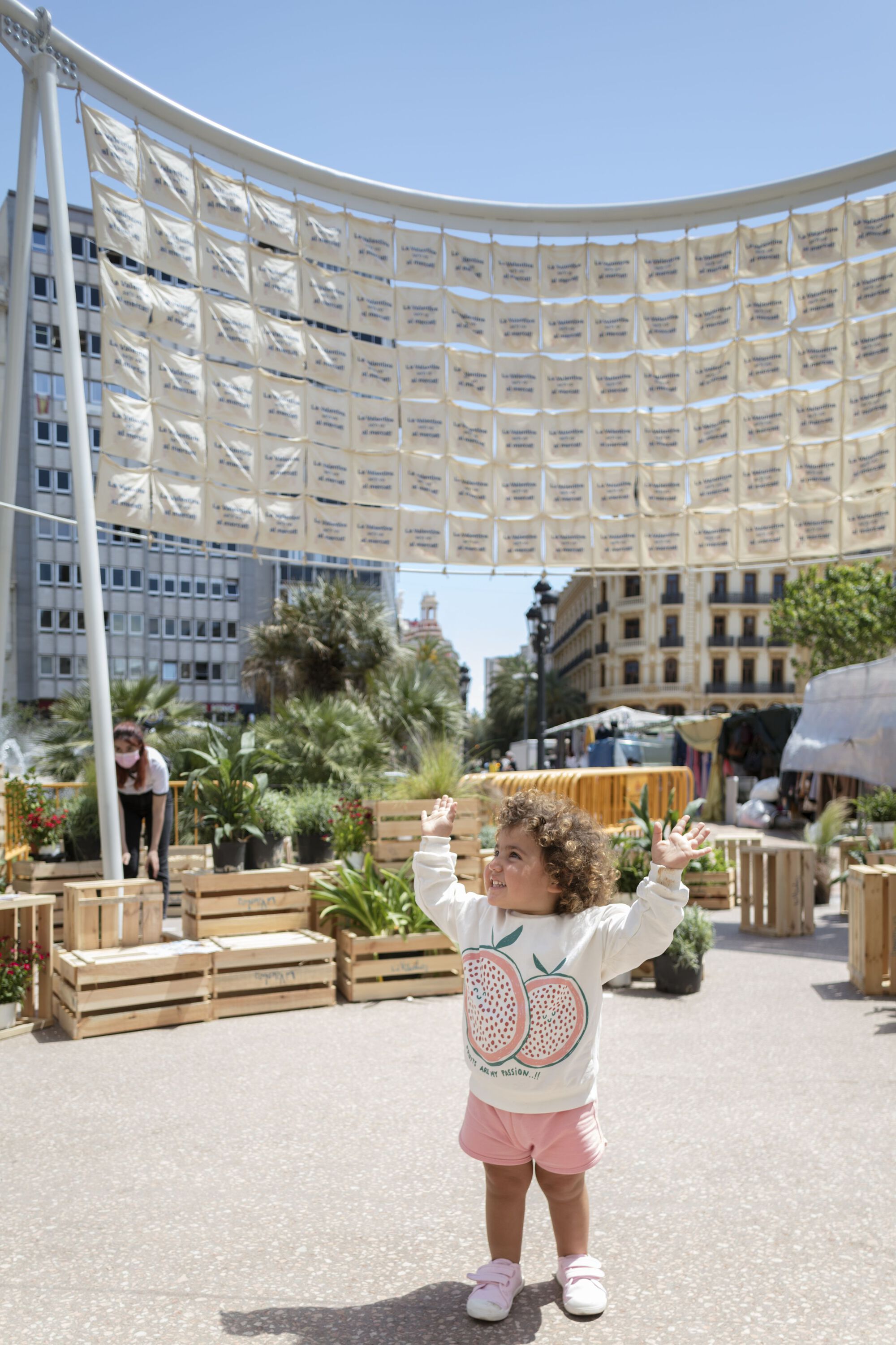 Child standing under the curved pavilion with timber furniture and planted beds in afternoon light