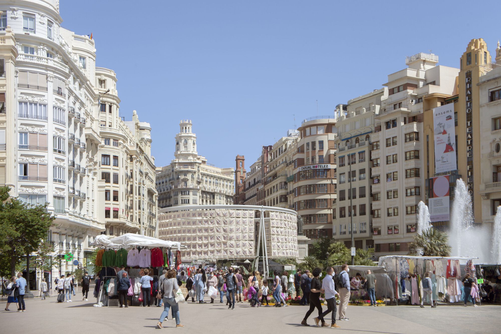 Temporary cylindrical pavilion with perforated white panels installed in a plaza surrounded by crowds and market stalls