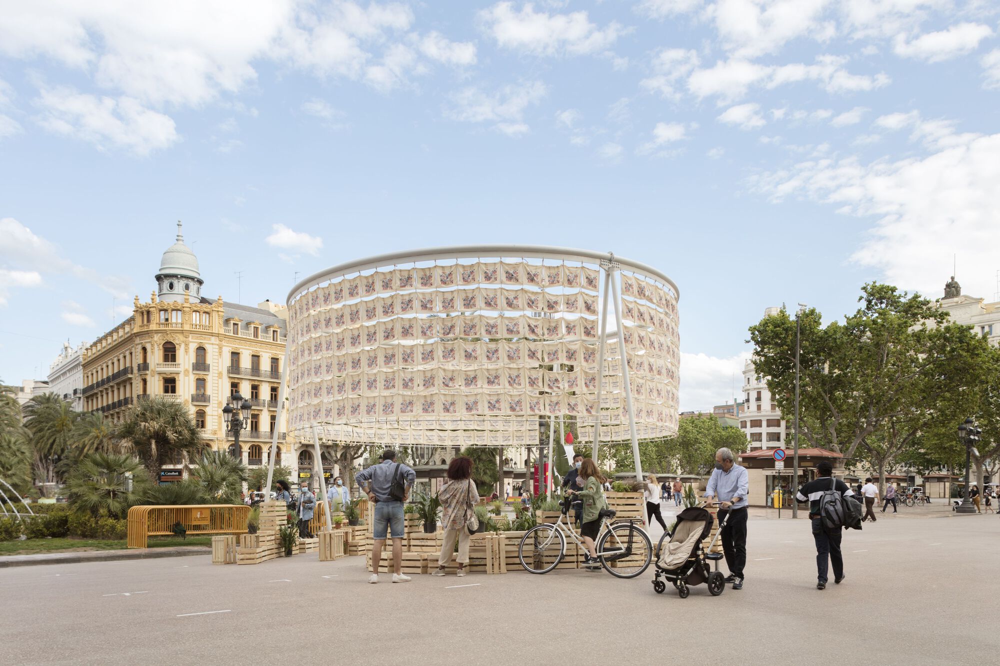 Cylindrical pavilion with patterned screen facade set in a public square with visitors and bicycles under sunny skies