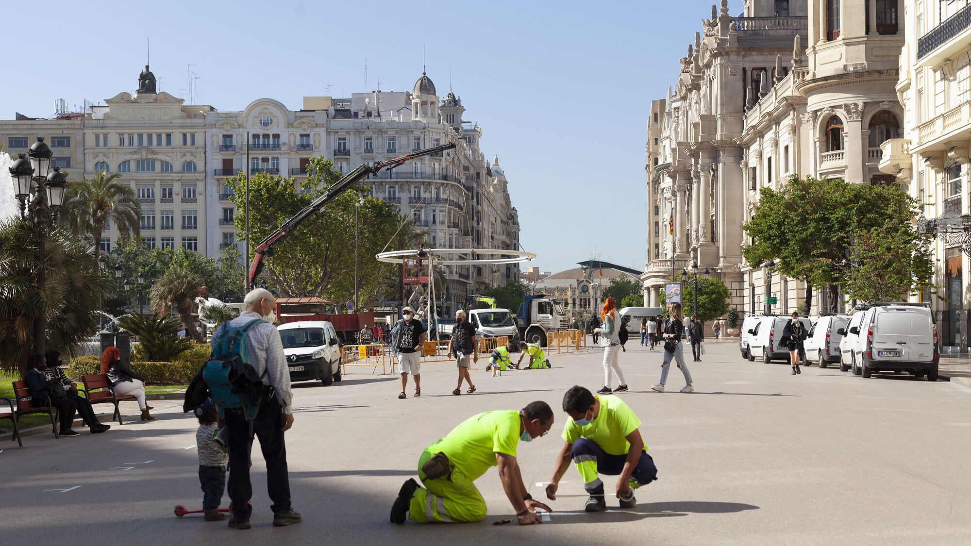 Construction crew measuring and marking positions on the plaza pavement as crane lifts structural element behind them