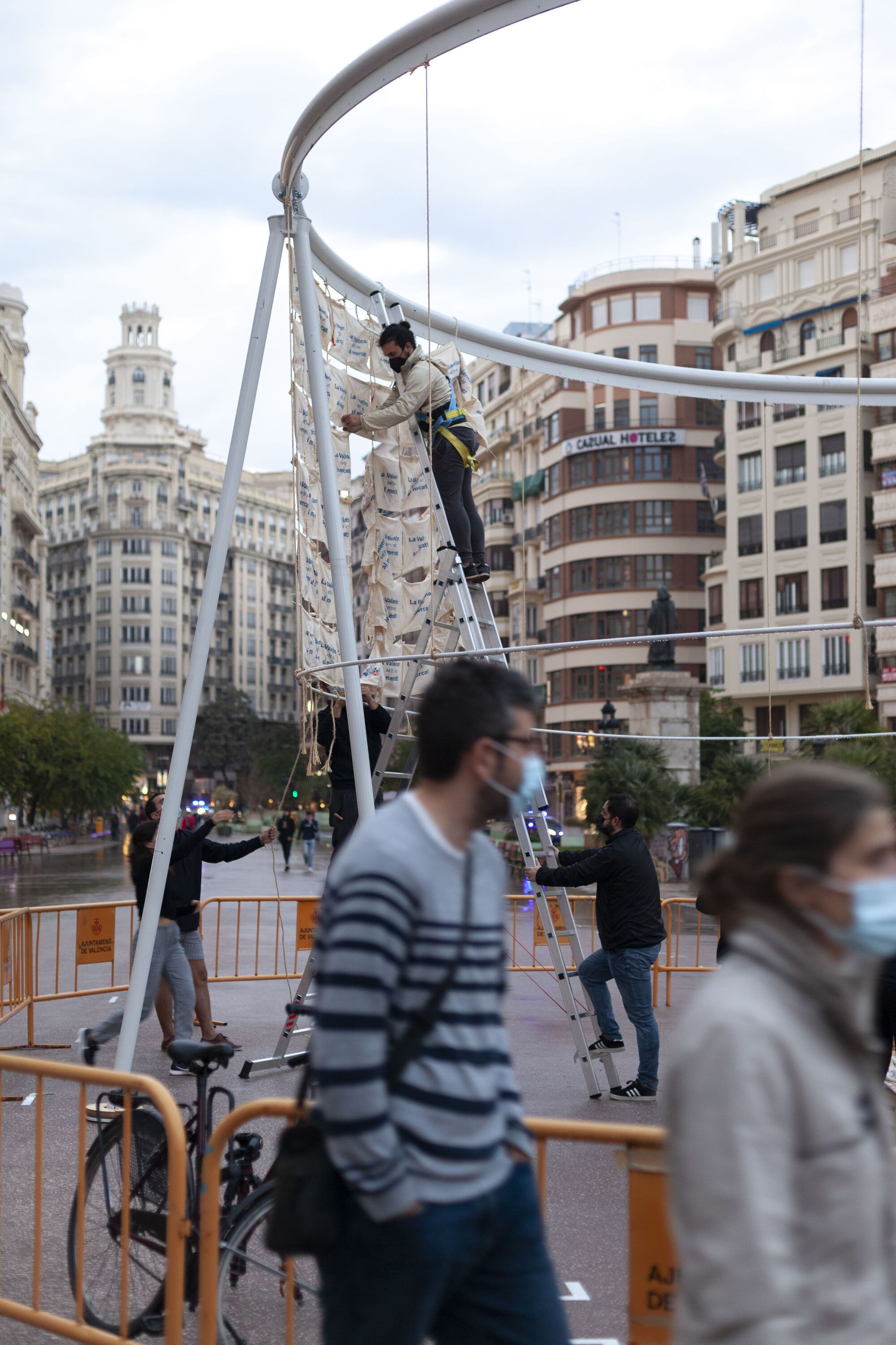 Temporary installation with curved metal frame and suspended fabric as pedestrians pass below wearing masks