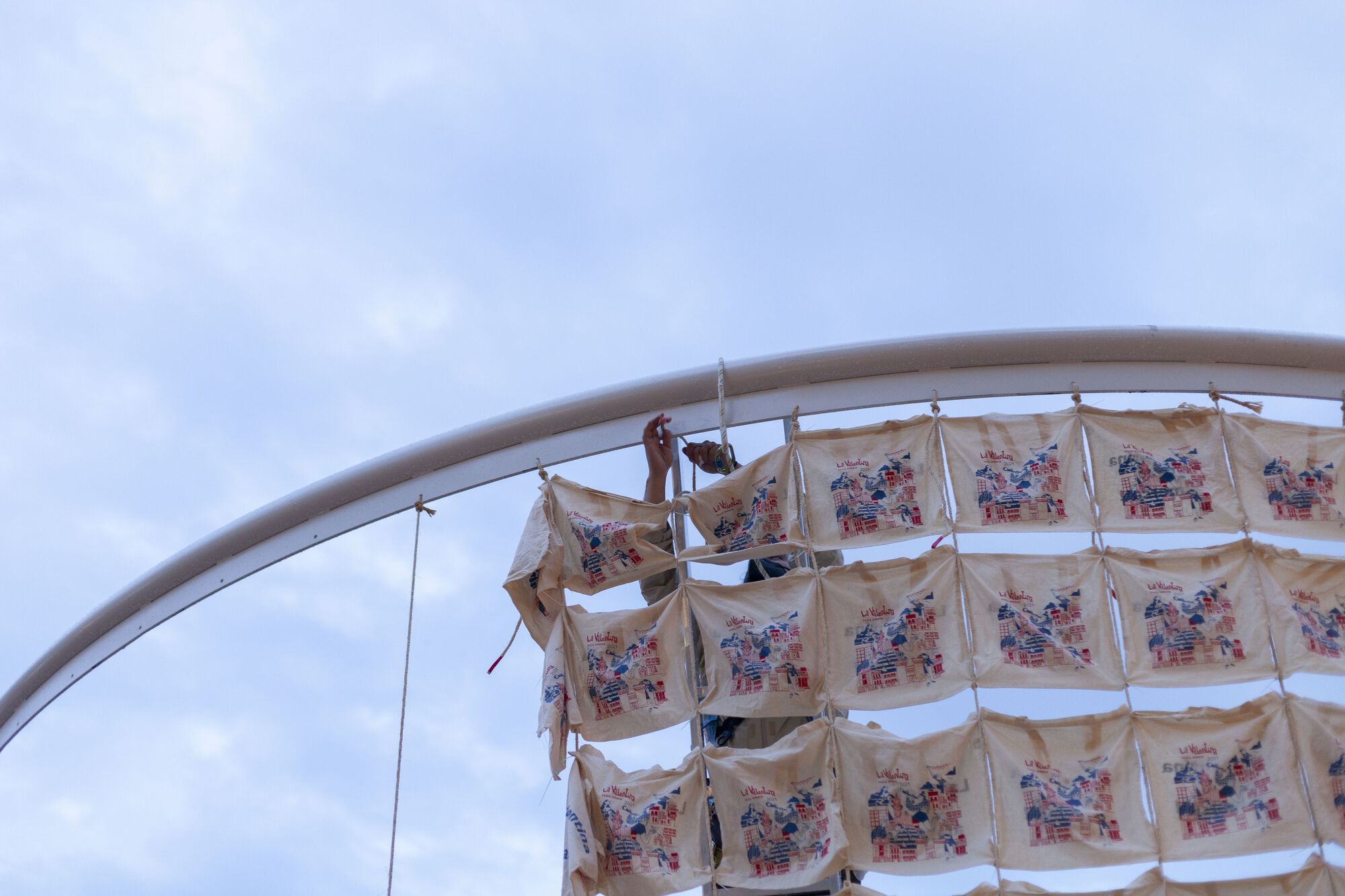 Close-up of curved steel armature with printed fabric banners suspended from cables against overcast sky