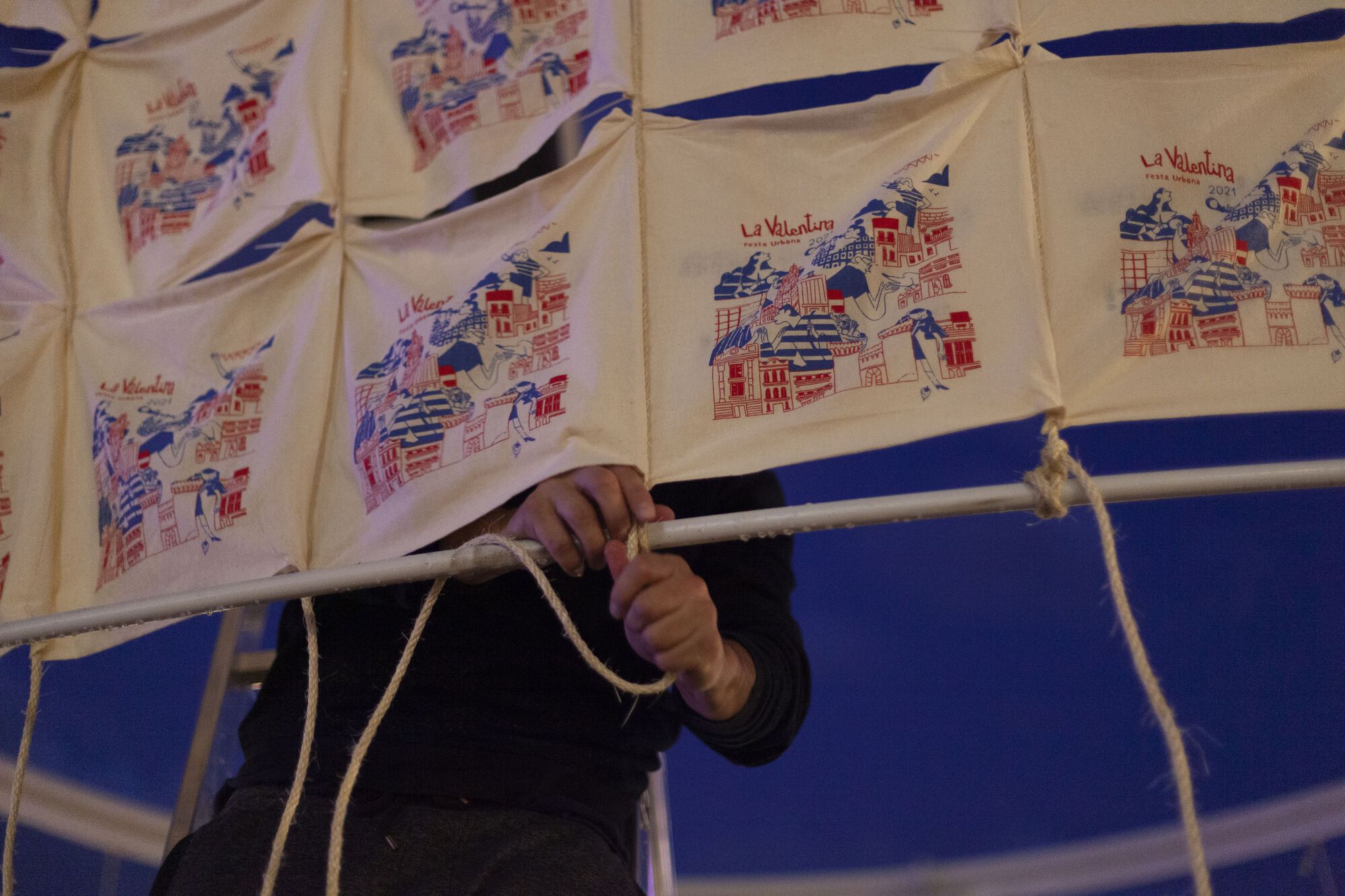 Hands tying rope knot on frame below printed textile panels at dusk