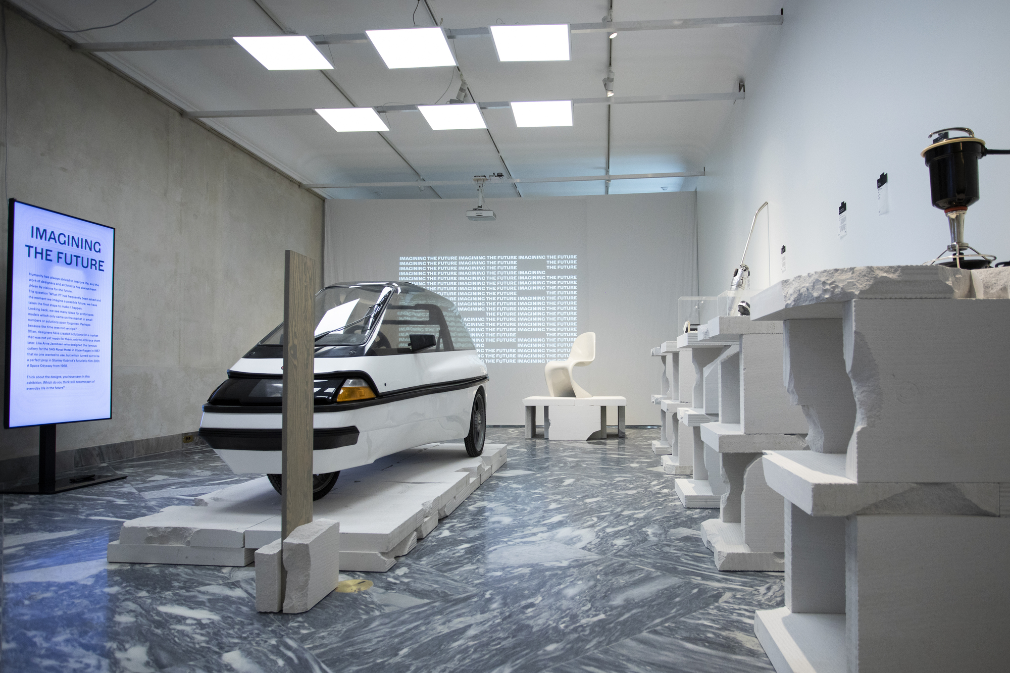 Gallery interior with concrete walls, suspended ceiling panels, and marbled grey flooring hosting sculptural objects