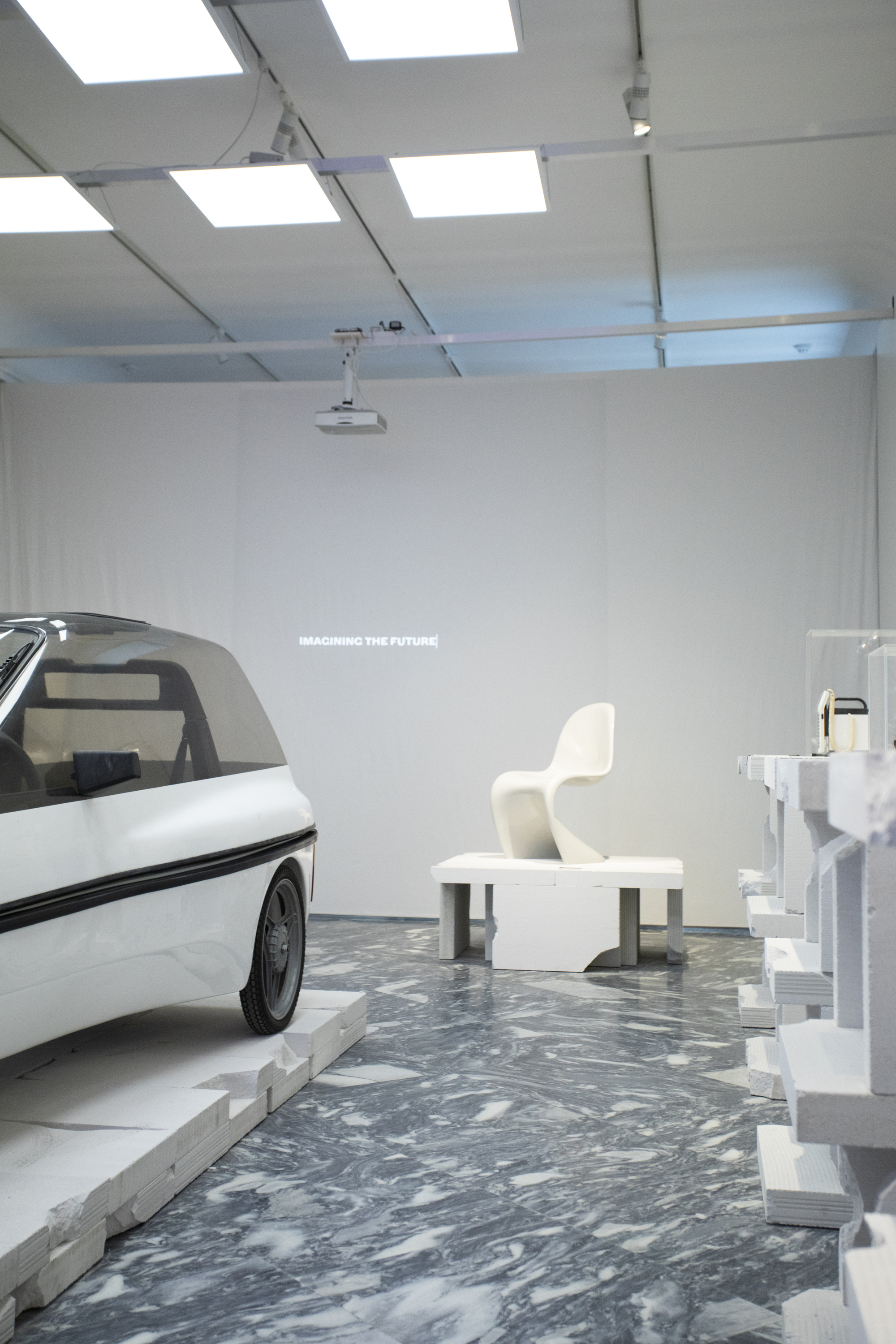 White exhibition room with gridded ceiling and marbled grey floor showing sculptural furniture arrangements