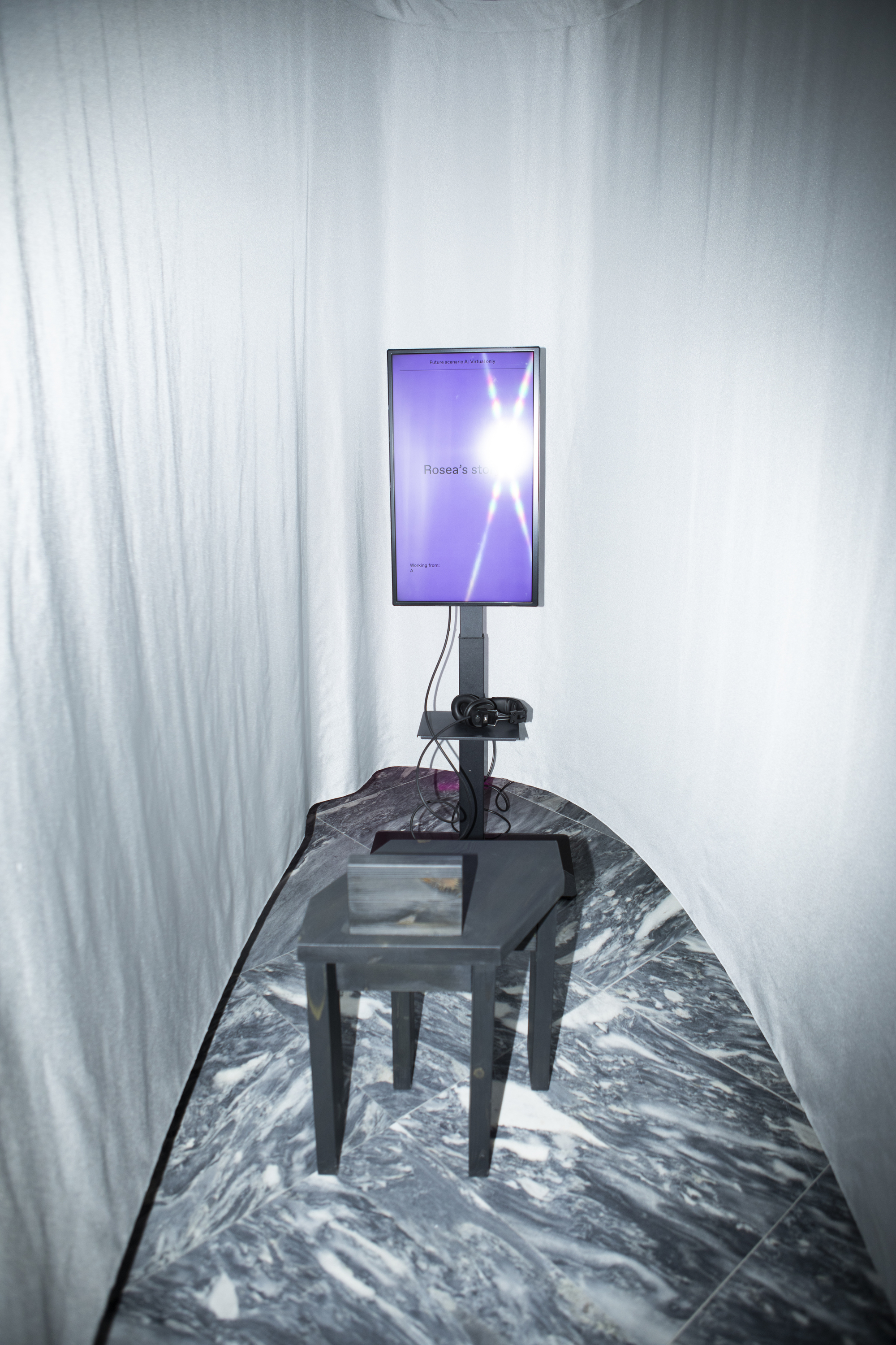 Digital screen on stand and glass table in narrow space lined with translucent curtains