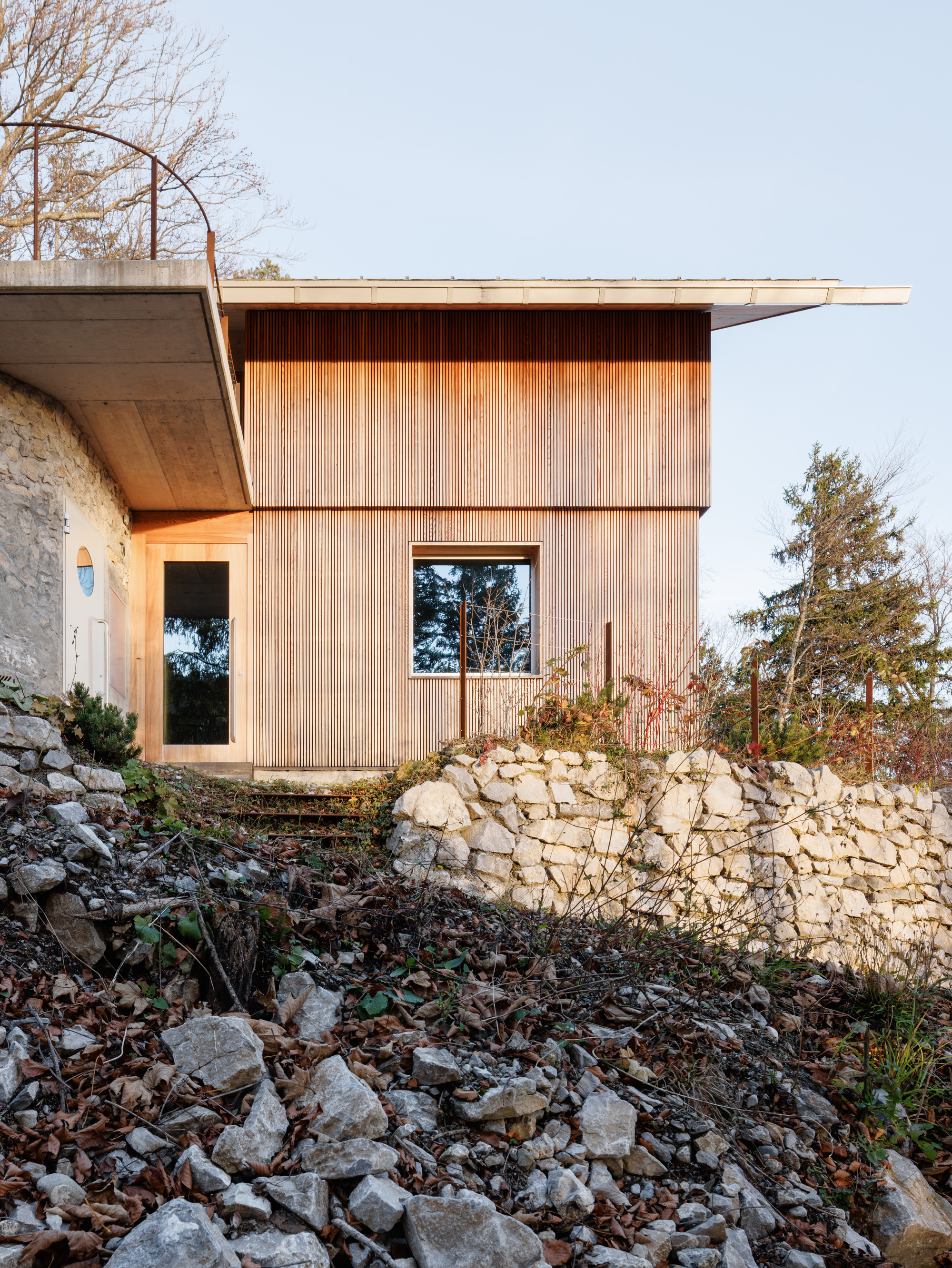 Vertical timber cladding with recessed window and concrete overhang above a stone retaining wall at dusk