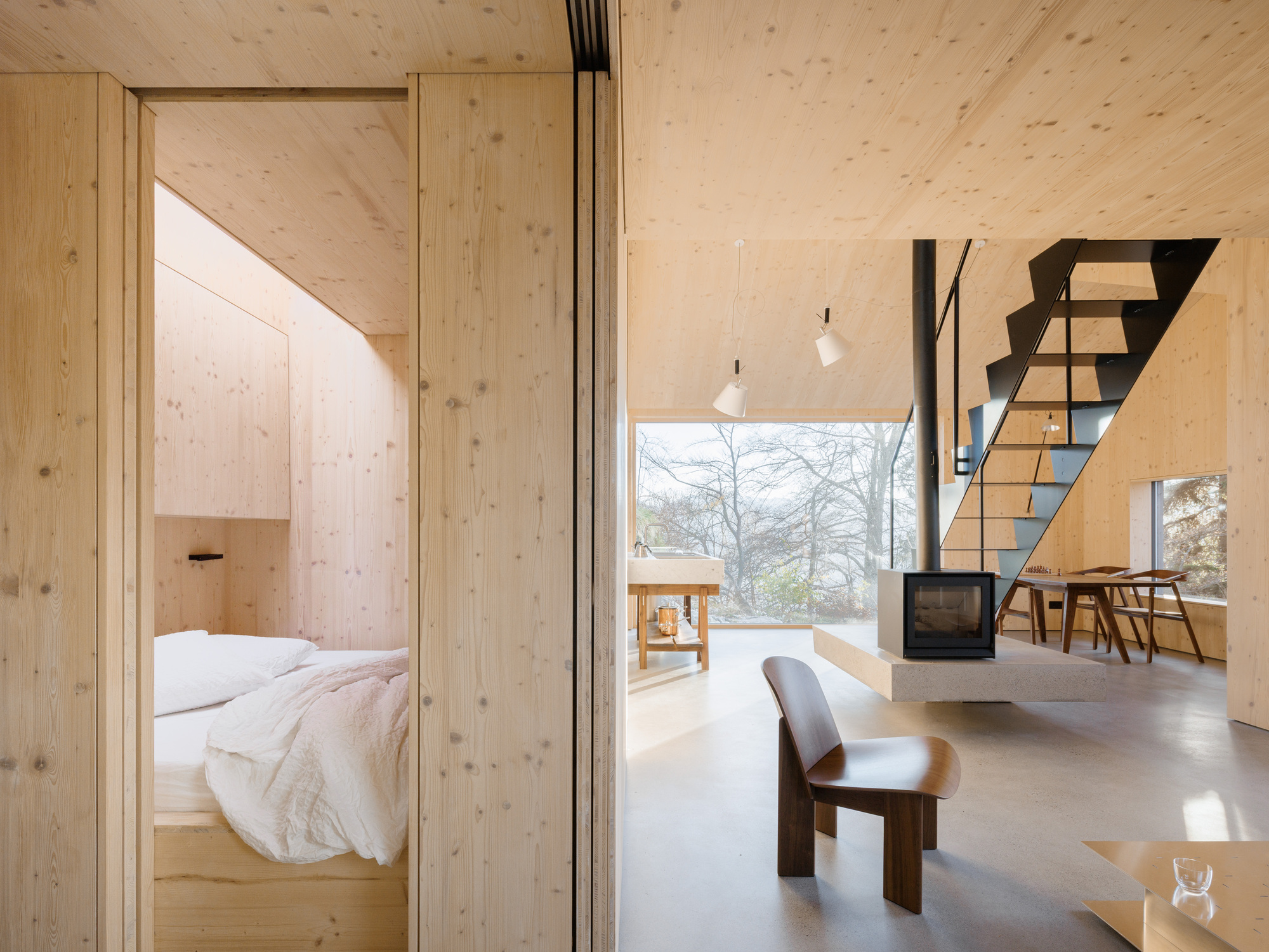 Open-plan interior showing bedroom alcove, cantilevered wood stove, and black steel staircase beside glazed wall